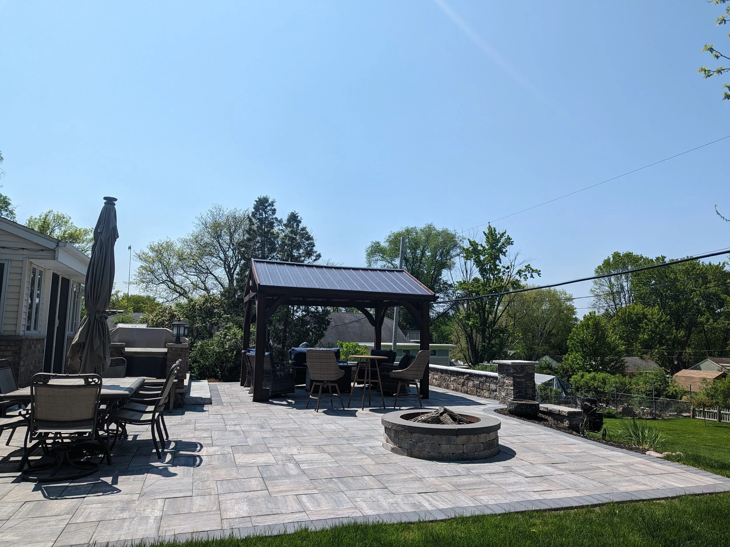 Backyard patio with outdoor furniture, a fire pit, pergola, umbrellas, and green trees under a clear blue sky.