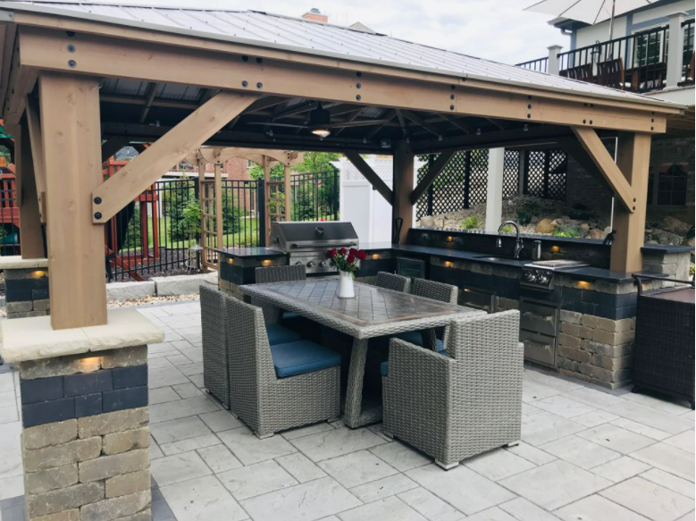 Outdoor covered patio with a dining table, wicker chairs, a built-in barbecue grill, and a vase with red flowers.