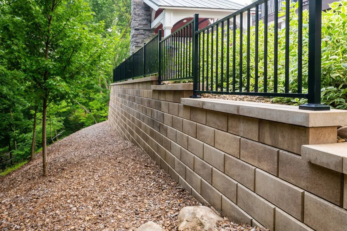 Large-format block retaining wall with powder-coated safety railing along a wooded slope — GoldGlo Landscapes