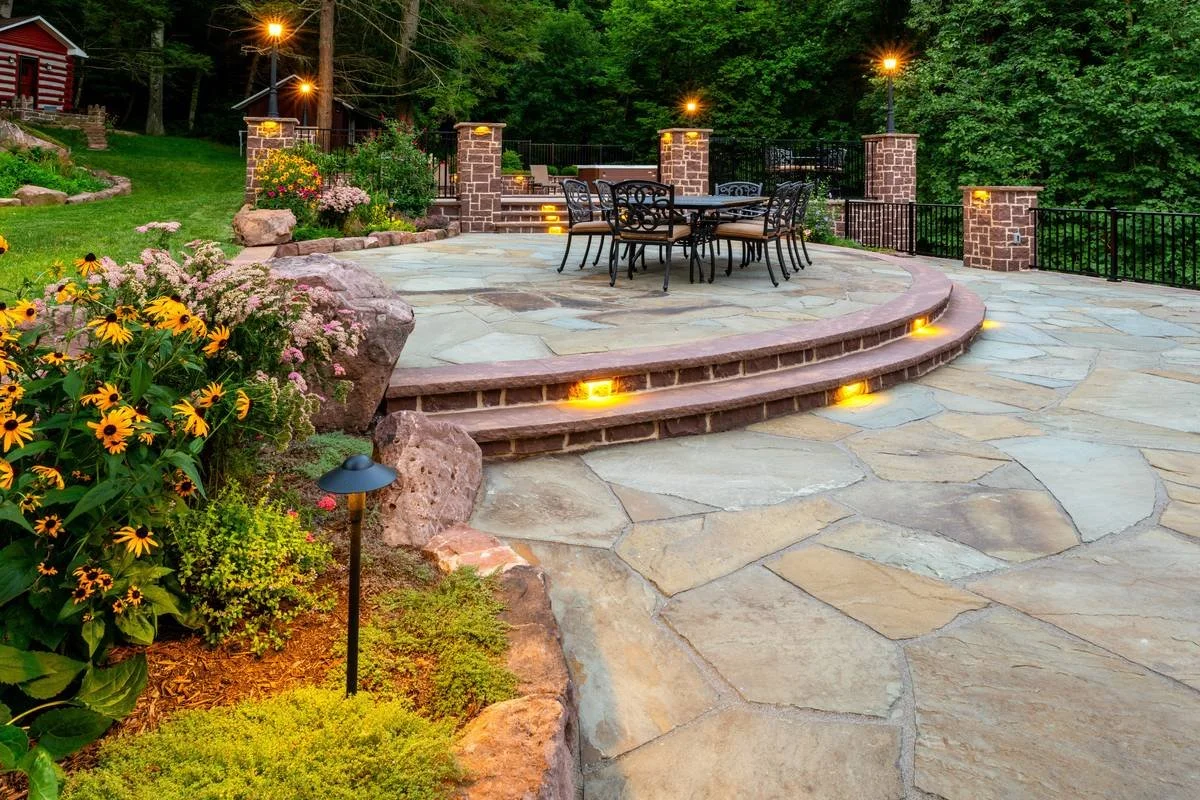 Lit brownstone steps rising to a raised flagstone dining area framed by stone columns — GoldGlo Landscapes