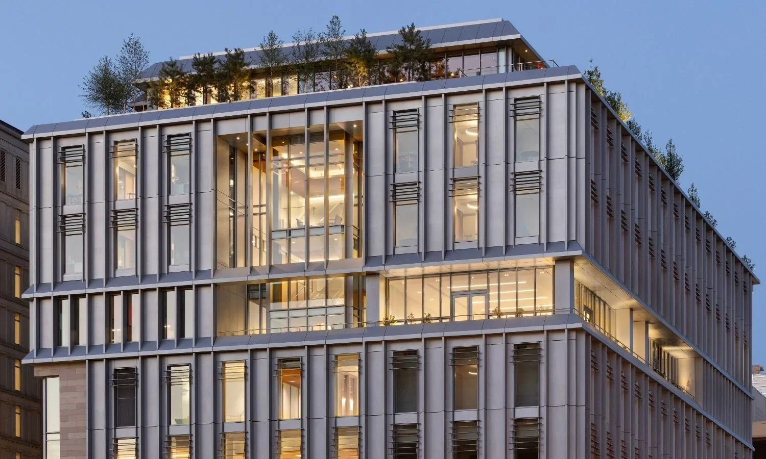 Evening view of Georgetown University’s McCourt School of Public Policy showing a modern glass-and-metal facade glowing with interior light, with trees and greenery in planters lining the illuminated rooftop terrace against a clear blue sky.