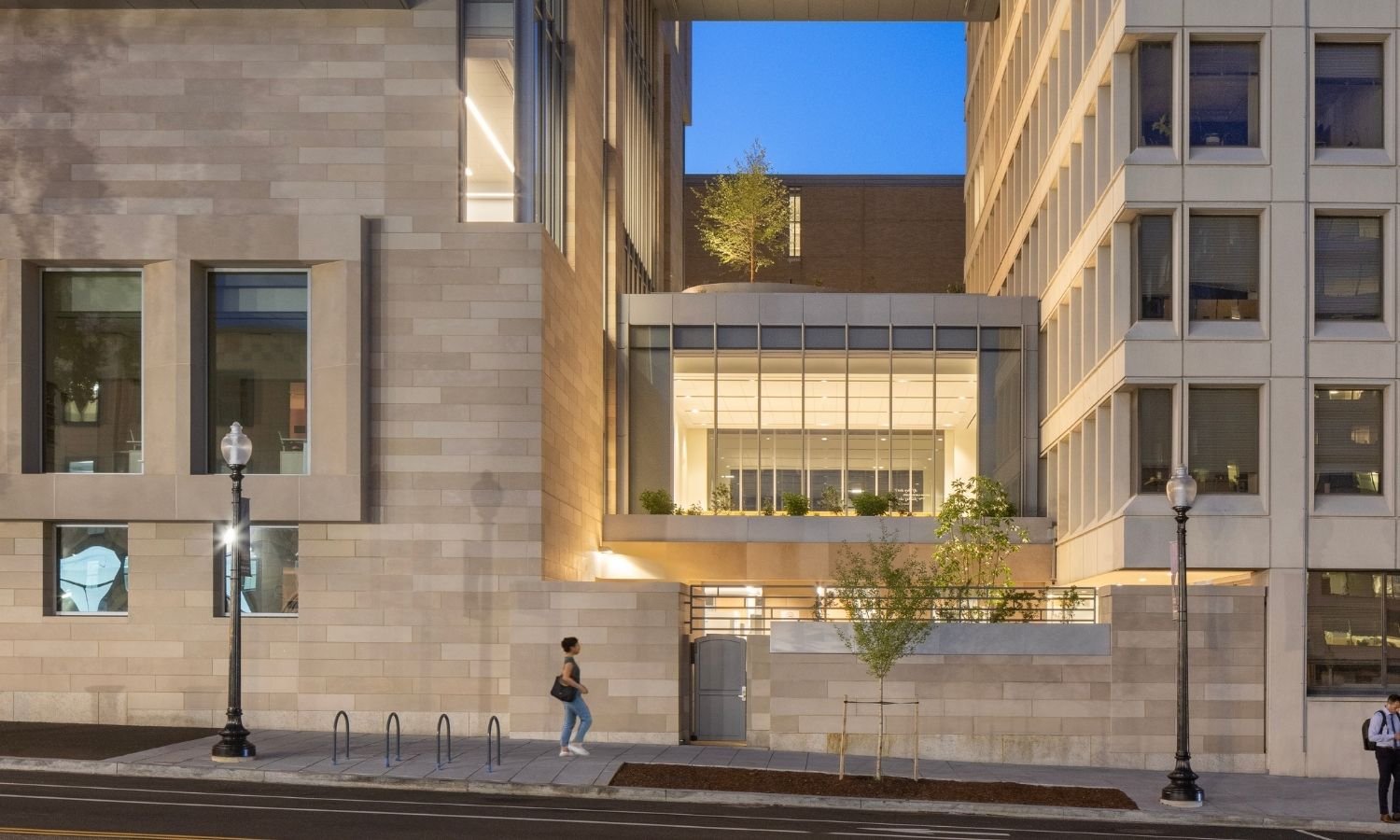 Dusk view of Georgetown University’s McCourt School of Public Policy showing a softly lit stone and glass facade with an elevated enclosed bridge connecting to a neighboring building, stacked terraces with planters and small trees.