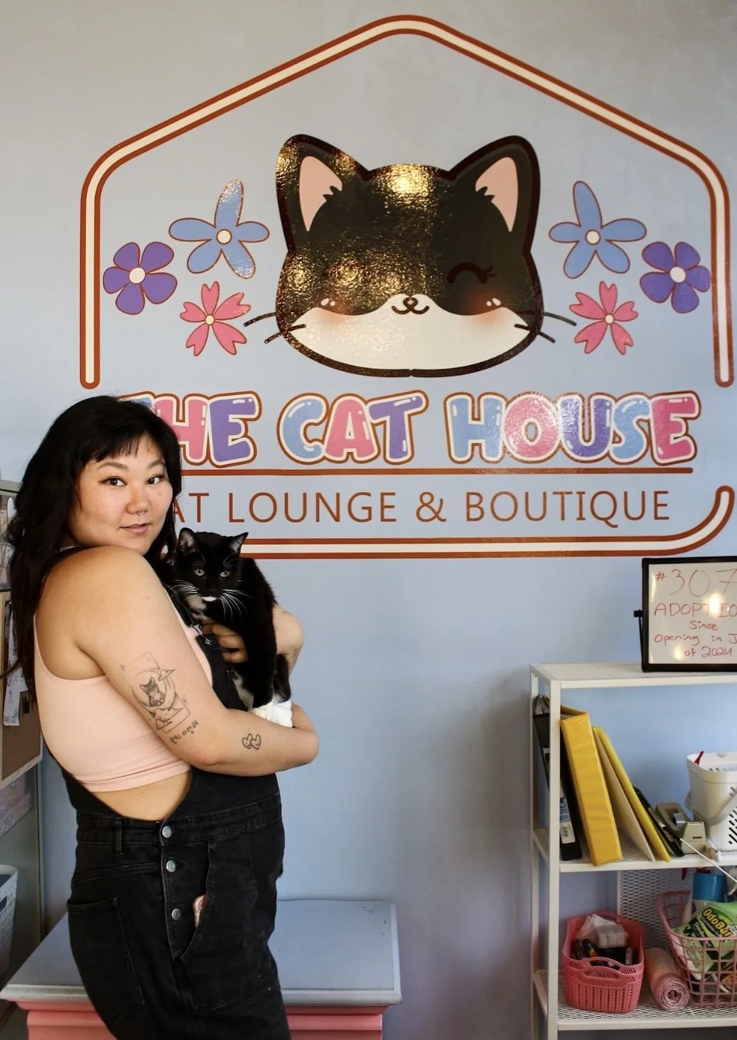Meet Irene, the woman that started The Cat House in front of a sign that reads 'The Cat House Cat Lounge & Boutique,' with a cartoon cat face and colorful flowers on the wall.