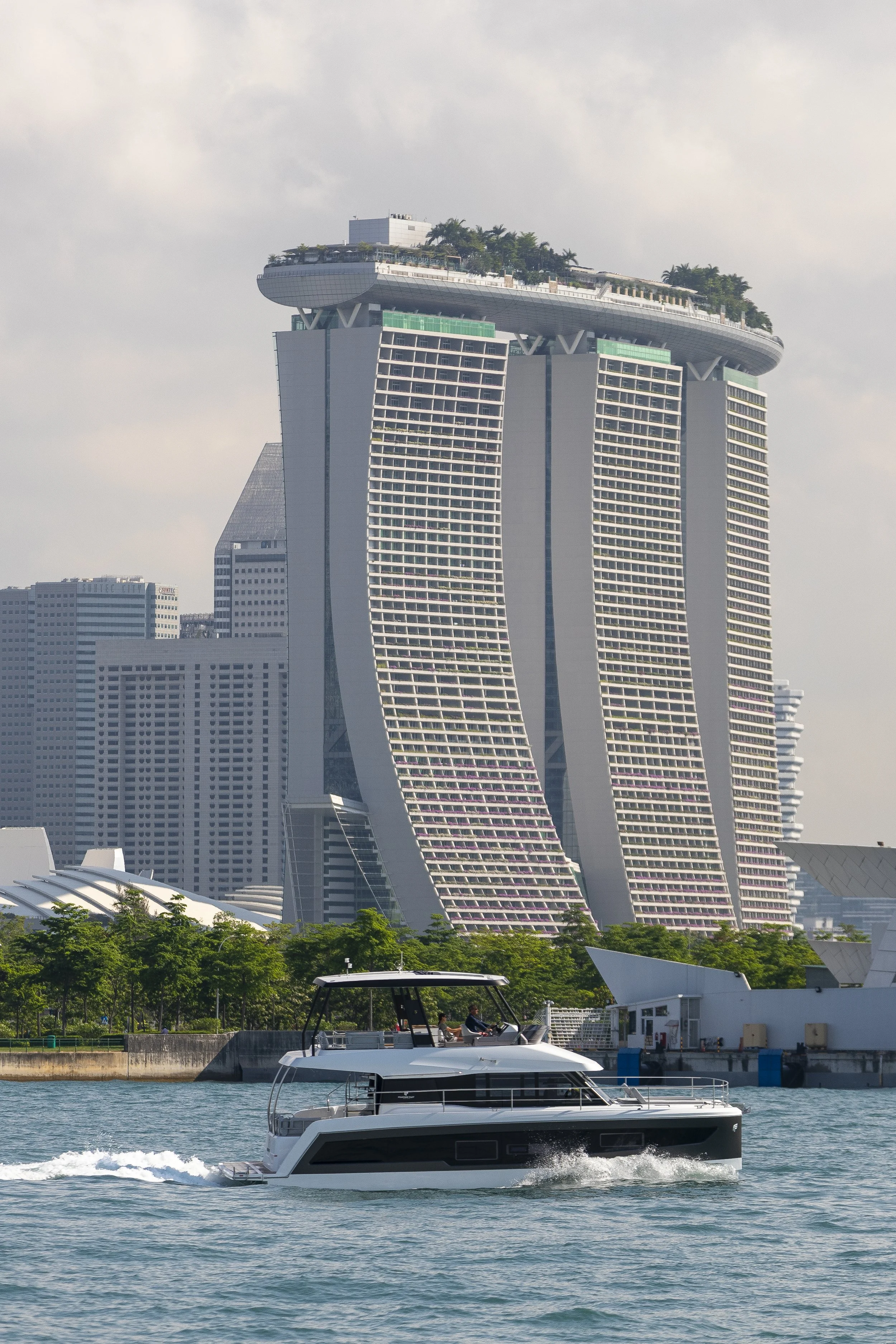 Modern skyscraper resembling a ship with three towers, lush greenery on the rooftop, and a luxury yacht cruising in the foreground.