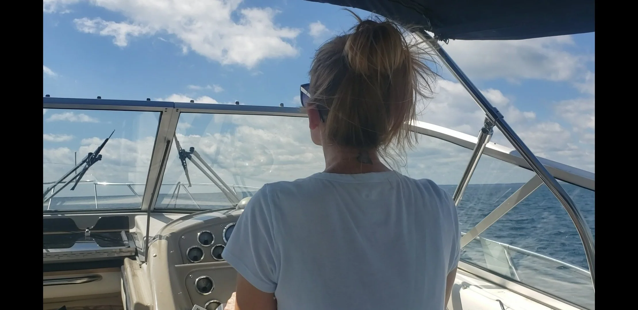 Person steering a boat on open water, sunny day with blue sky and clouds, rear view