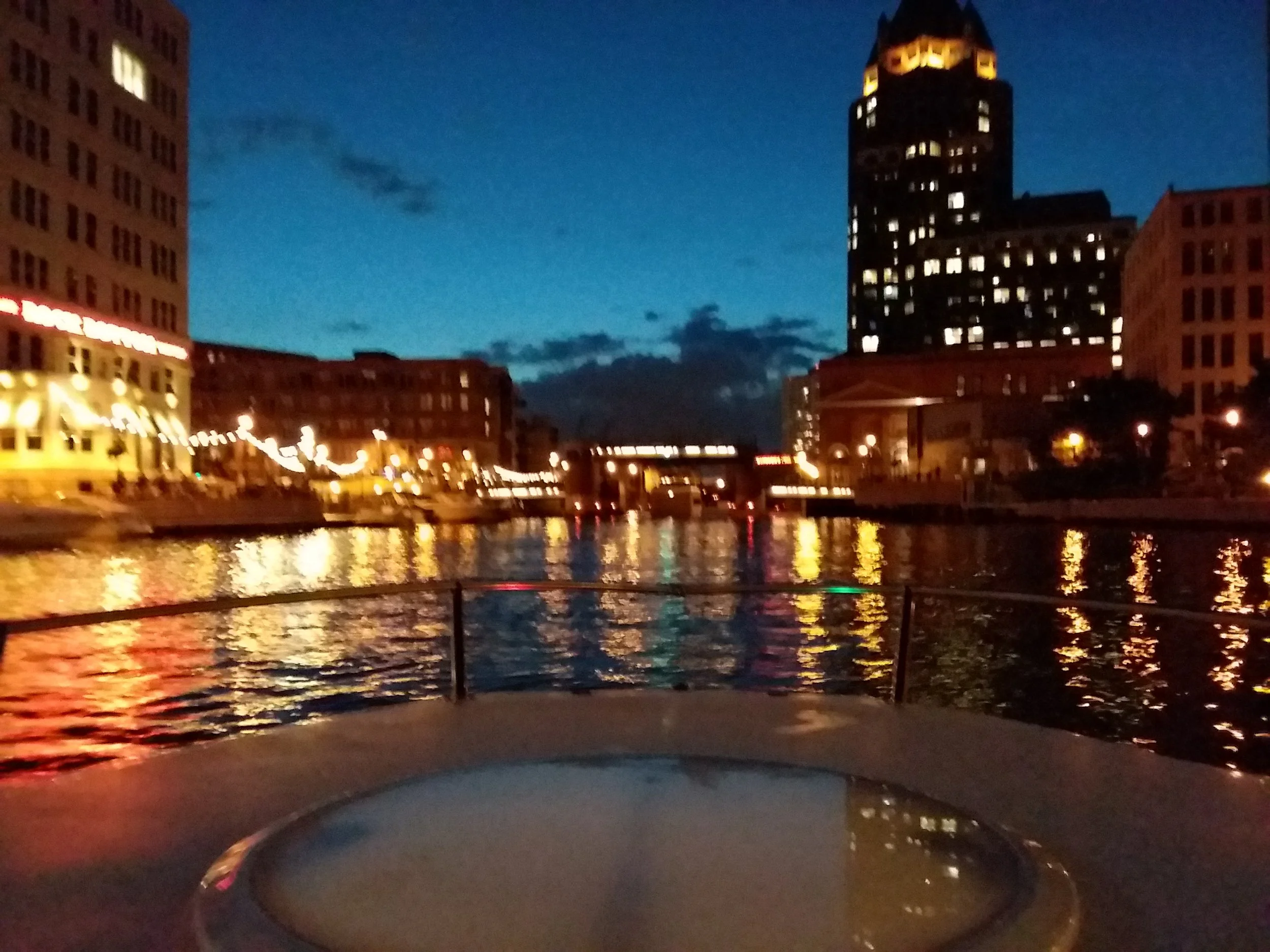 City skyline at dusk with illuminated buildings, river reflection, and boats along the shoreline.