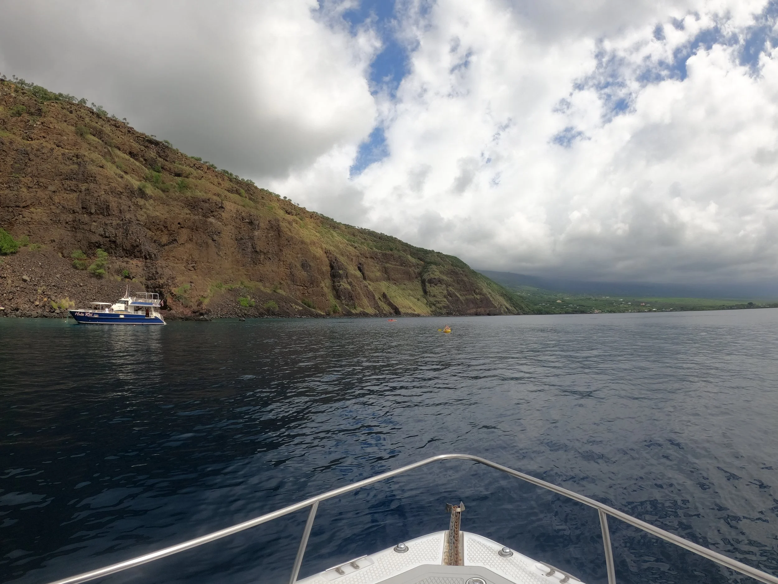 Perspective from a boat on the water, with a rocky cliffside and another boat in the distance, under a cloudy sky.