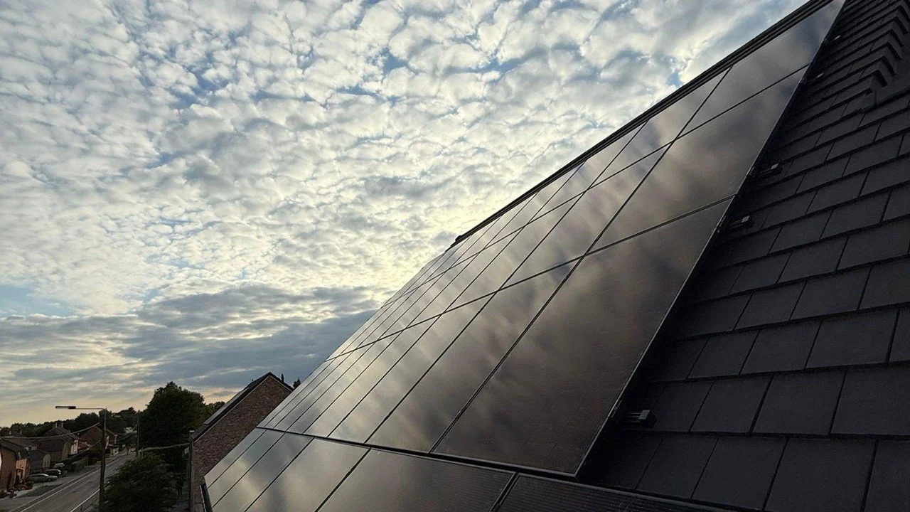 Zwarte zonnepaneel op een dak, met een bewolkte lucht die weerspiegelt in de panelen.