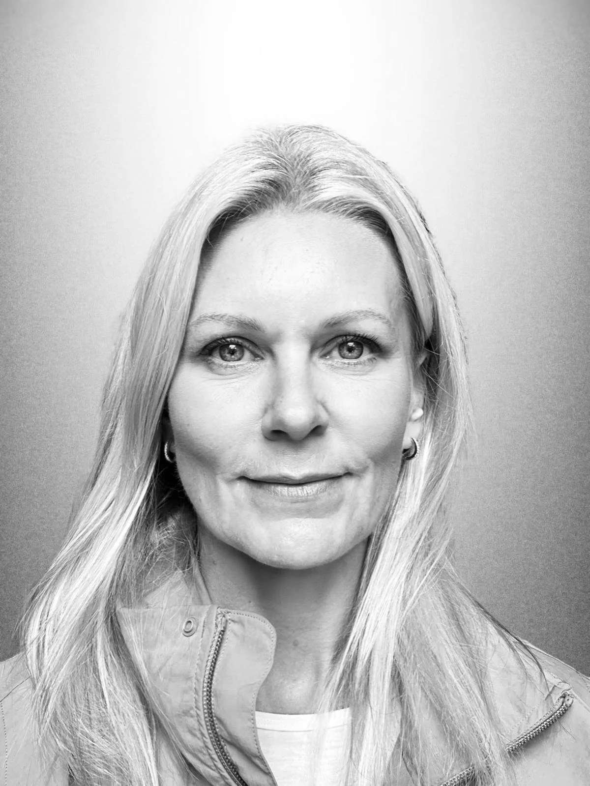 A smiling woman with light hair pulled back, wearing a tank top and a delicate necklace, standing outdoors in front of a brick wall.