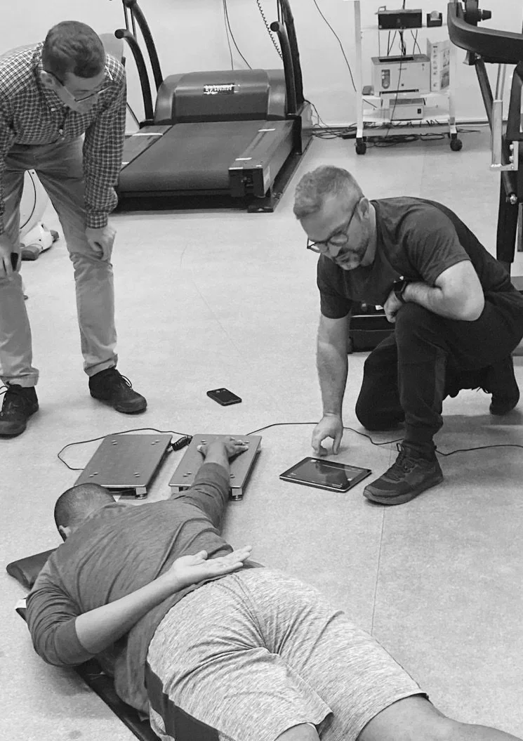 A person lying on a mat in a medical or physical therapy setting, with two other individuals assisting or observing, and various electronic equipment and a treadmill in the background.