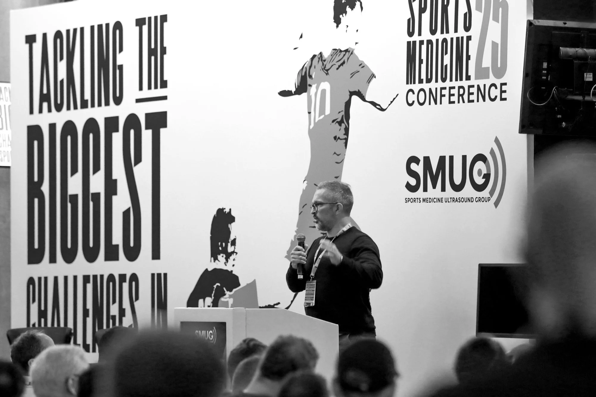 A man speaking into a microphone at a conference with a large poster behind him that reads 'TACKLING THE BIGGEST CHALLENGERS IN SPORTS MEDICINE.' The poster includes the logos for the Sports Medicine Ultrasound Group and the annual conference. The man stands behind a podium with a badge hanging around his neck, addressing an audience.