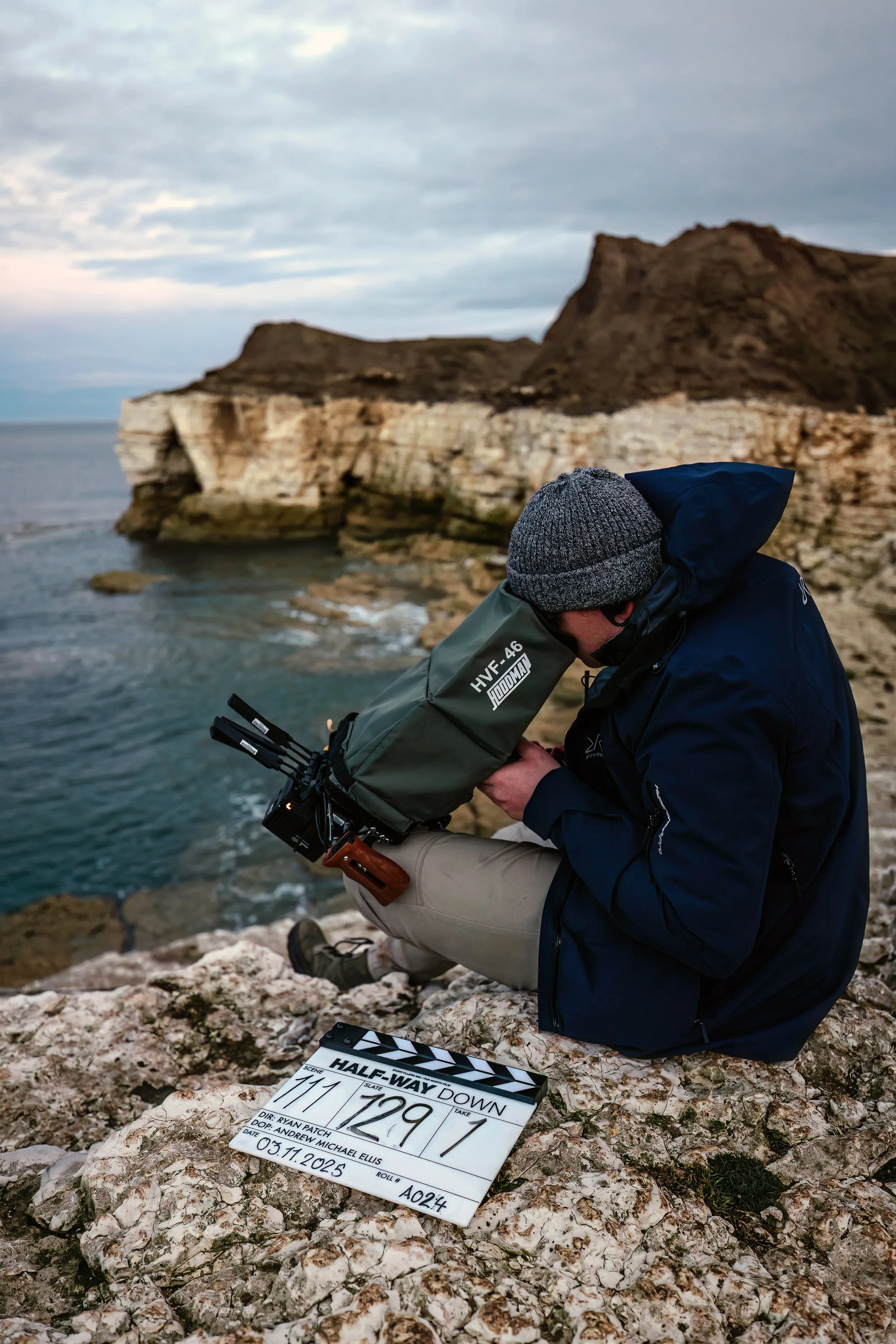 A man in outdoor clothing, wearing a gray knit hat, is sitting on rocky terrain whilst focus pulling, with a close-up of a film slate on the ground in front of him. The background shows a rugged coastline with cliffs, ocean, and a cloudy sky.