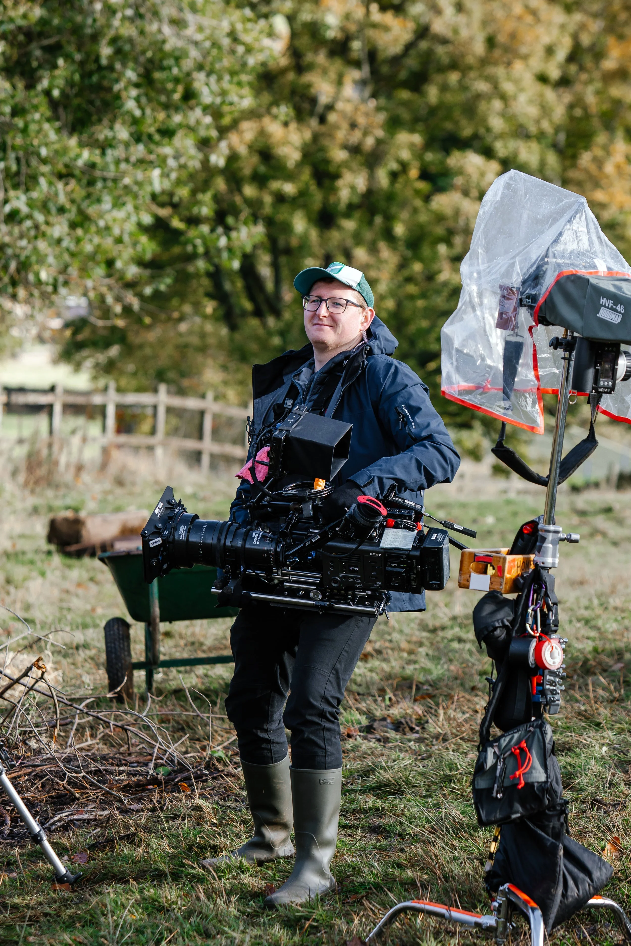 A man standing outdoors in a grassy area with trees in the background, holding a professional film camera. He wears a green cap, glasses, a navy jacket, gray pants, and gray rubber boots. There is professional filming equipment nearby, including a large light with a transparent cover and a black and red bag on the ground.
