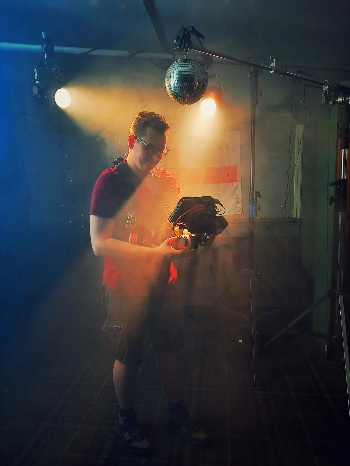 Person standing in a dimly lit room holding a camera, with a disco ball overhead and stage lights shining.