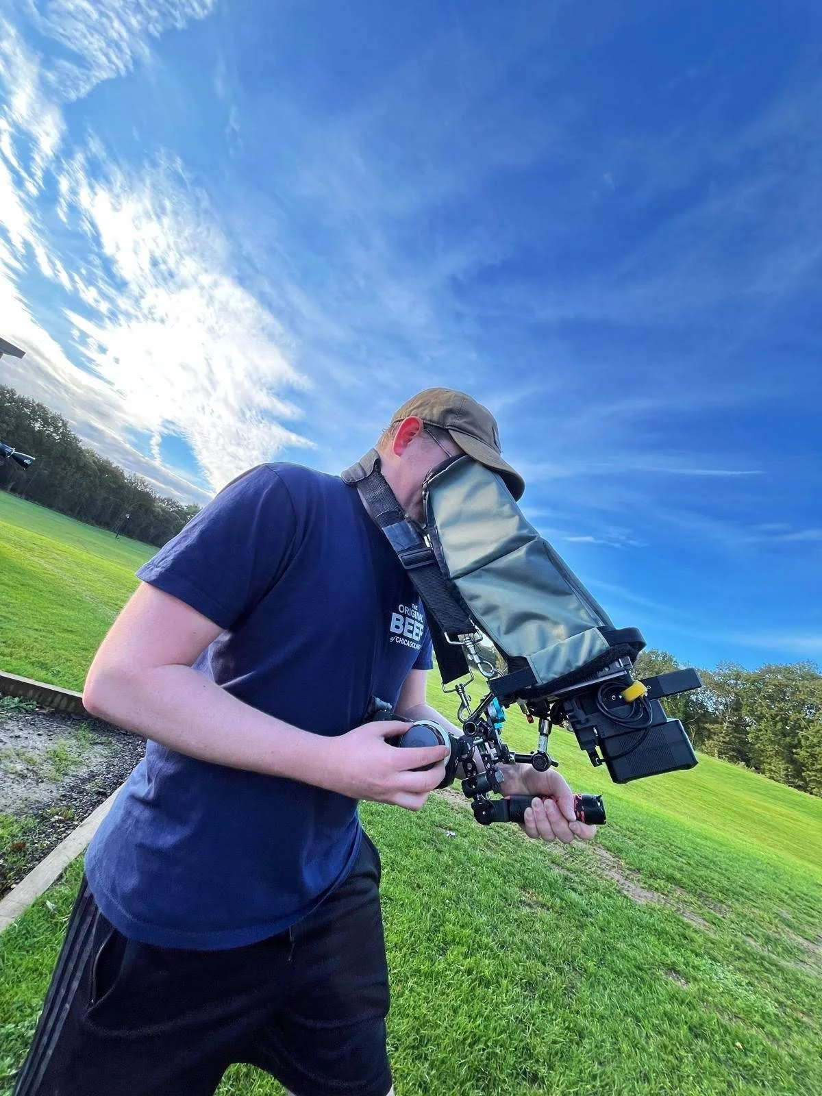 A person operating a focus pulling rig outdoors on a grassy field with trees and a partly cloudy sky in the background.