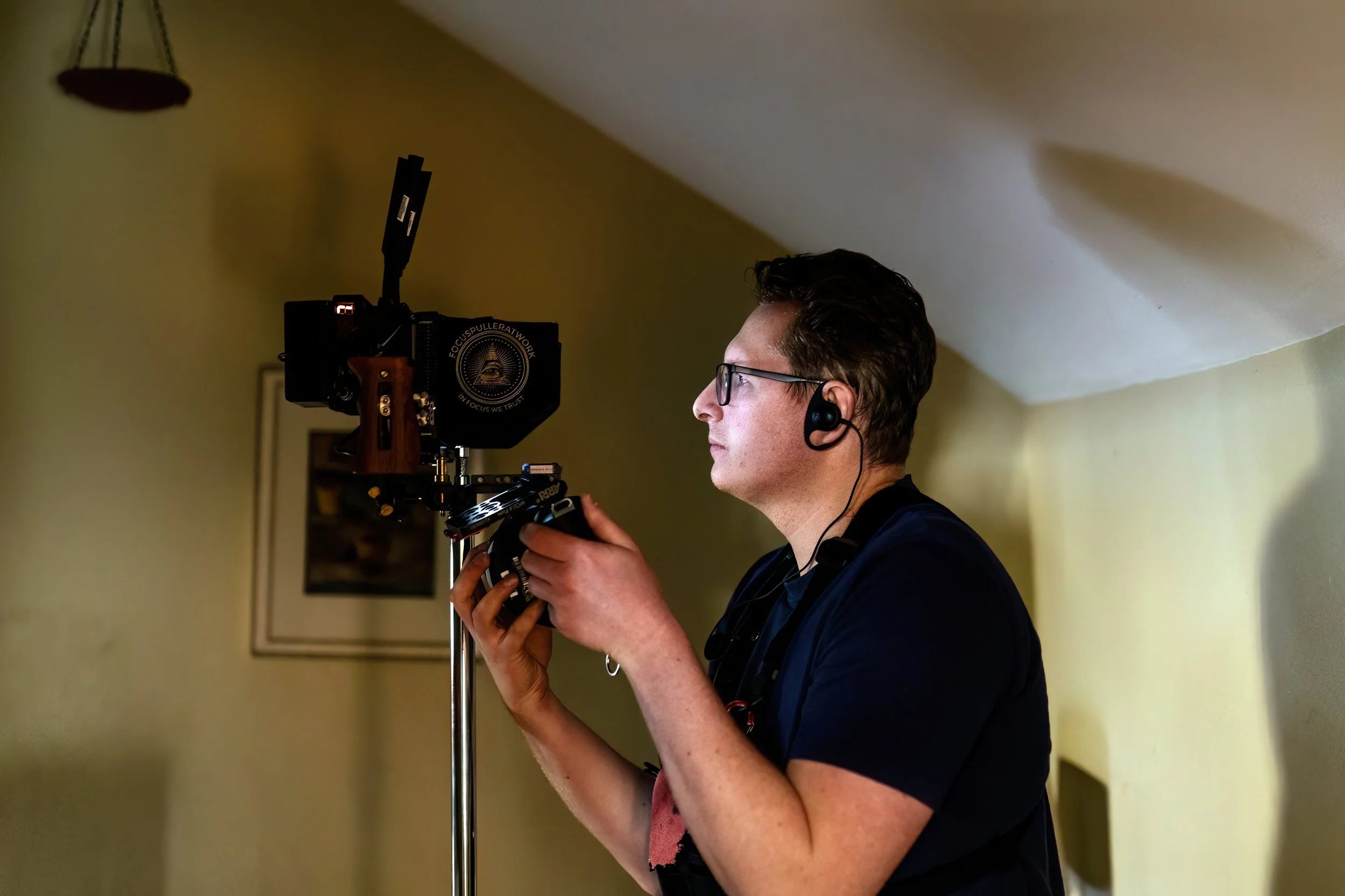 A person with short dark hair, glasses, and a black shirt is focus pulling intently, wearing an earpiece, in a room with a sloped ceiling and a framed picture on the wall.