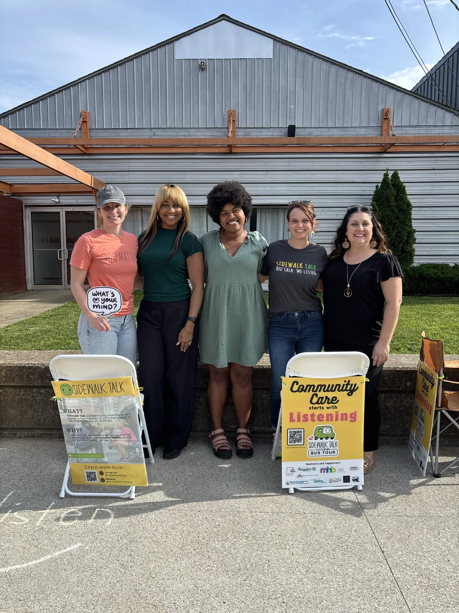 Five women standing side by side outdoors in front of a building, promoting a community listening event with signs that say "Sidewalk Talk" and "Community Care starts with Listening."