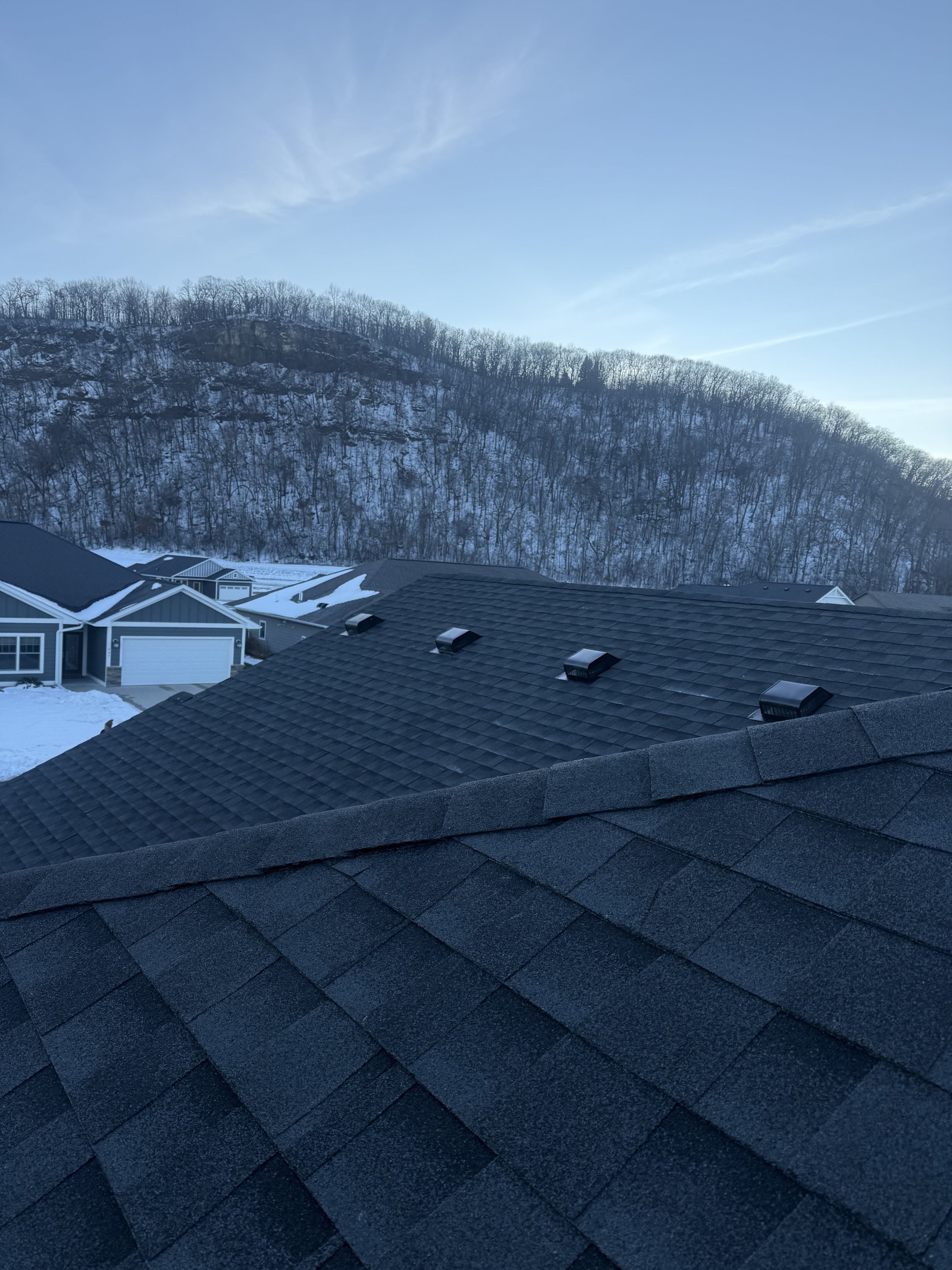 A dark gray roof that was installed in Lake City, MN by Skyline Roofing. The background features additional homes in the neighborhood and the scenic bluffs of SE MN.