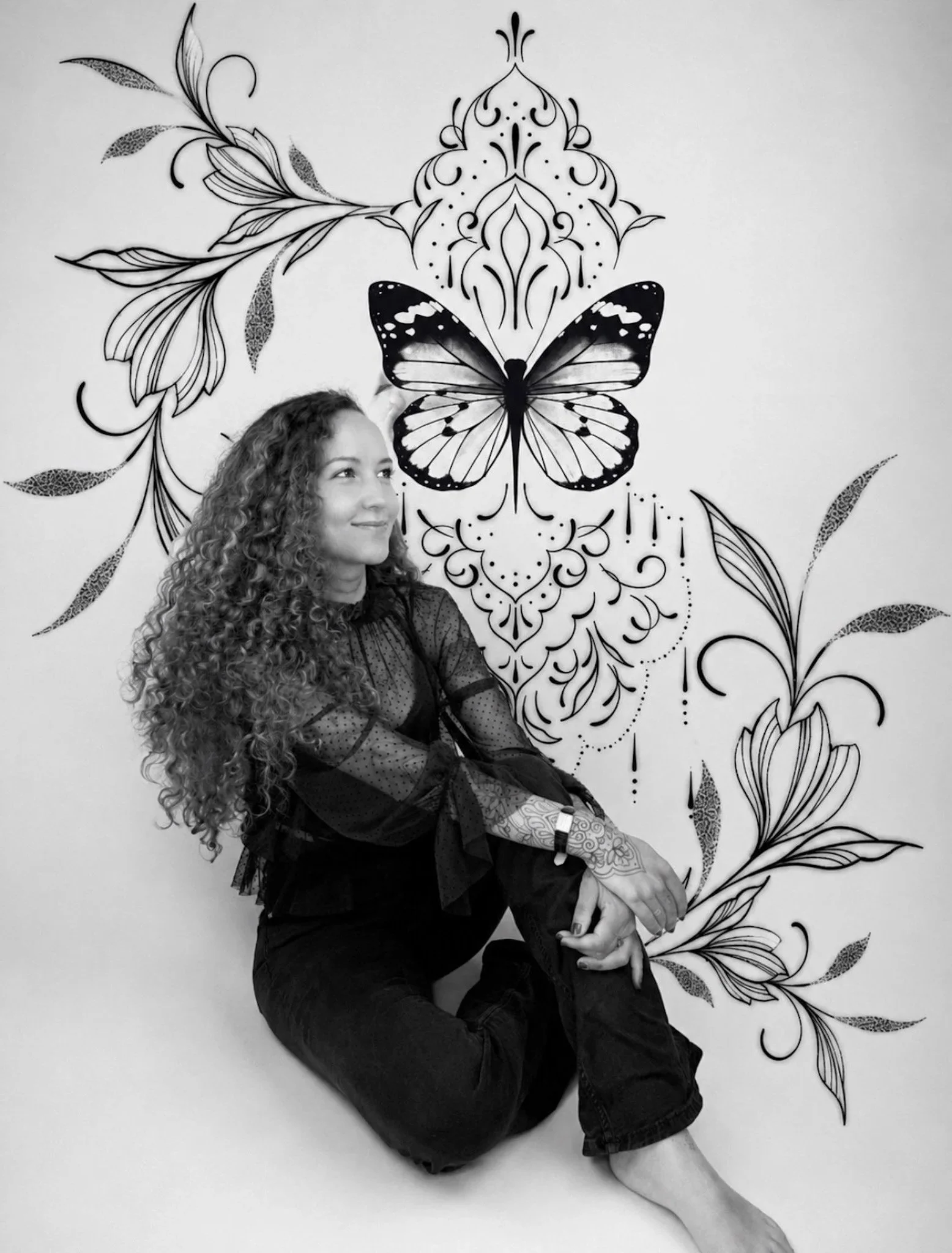 A woman with curly hair sitting against a white wall with a decorative black and white butterfly and floral design.