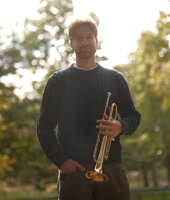 A man with red hair and a floral shirt playing a trumpet in front of a piano with wooden paneling.