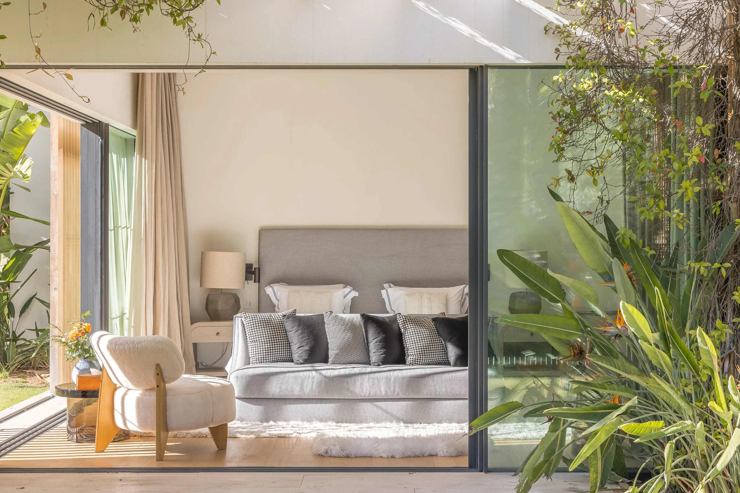Modern living room with large glass sliding doors, view of courtyard garden with lush green plants, beige curtains, gray upholstered bed with pillows, side table with lamp, armchair, and rug.