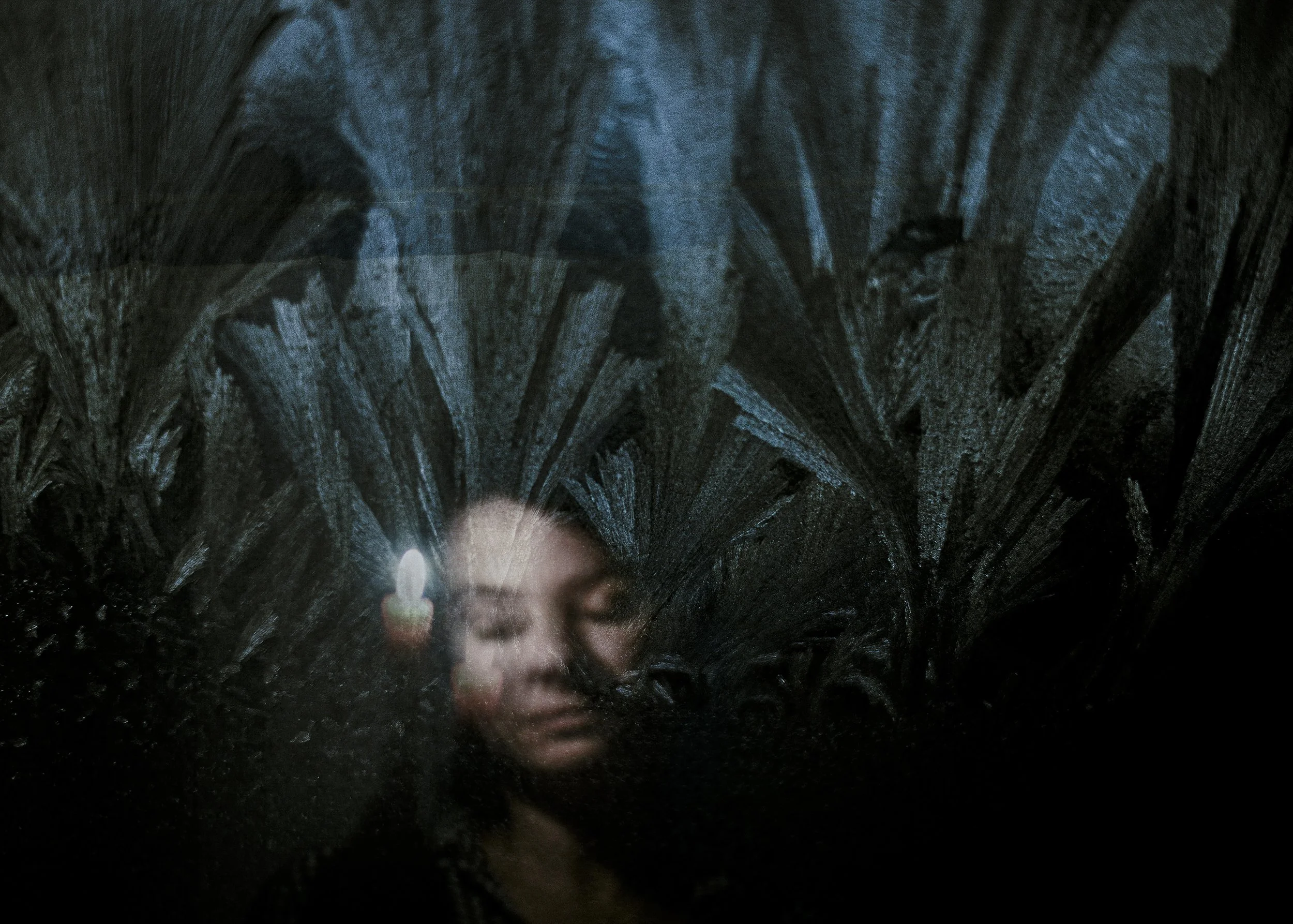 Close-up of a woman with closed eyes behind a glass window with dark, textured patterns, and a candle reflection.