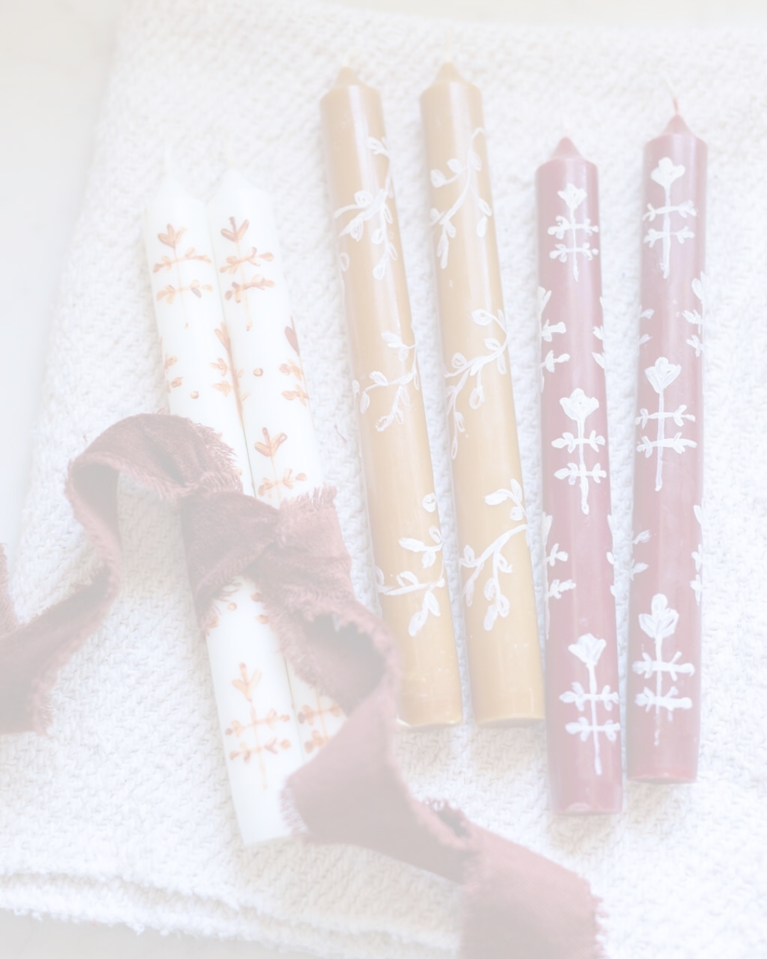 Six decorative candles with white floral patterns, two gold, two pink, and two white, sitting on a white textured cloth with a brown ribbon nearby.