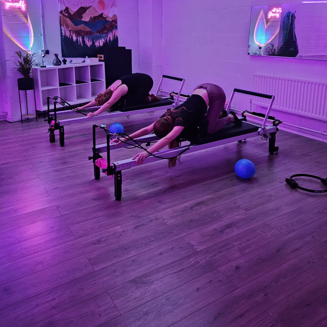 Three people practicing yoga on reformer Pilates machines in a studio with beige curtains.