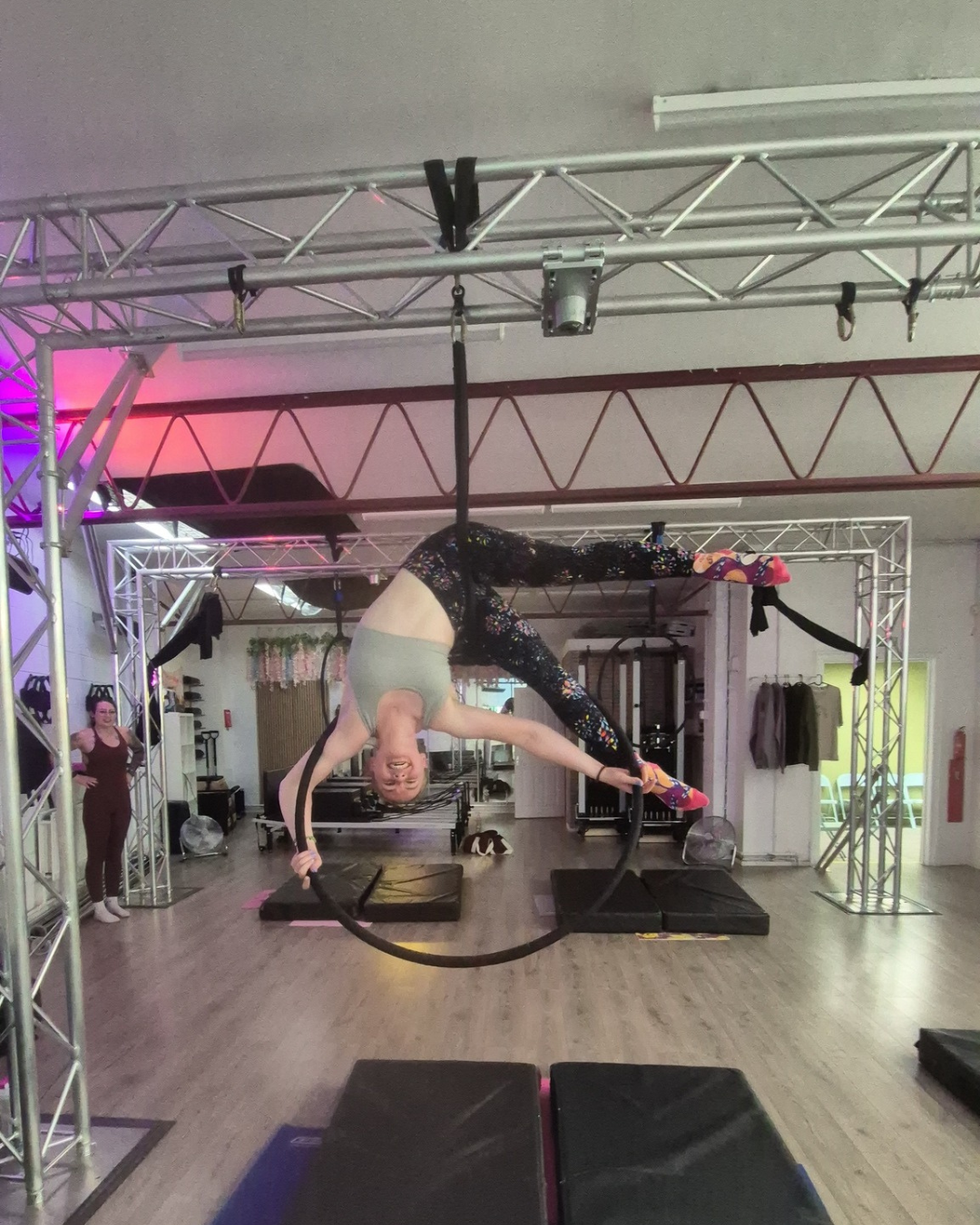 A woman performing an aerial acrobatic move on a black aerial hoop against a plain background.