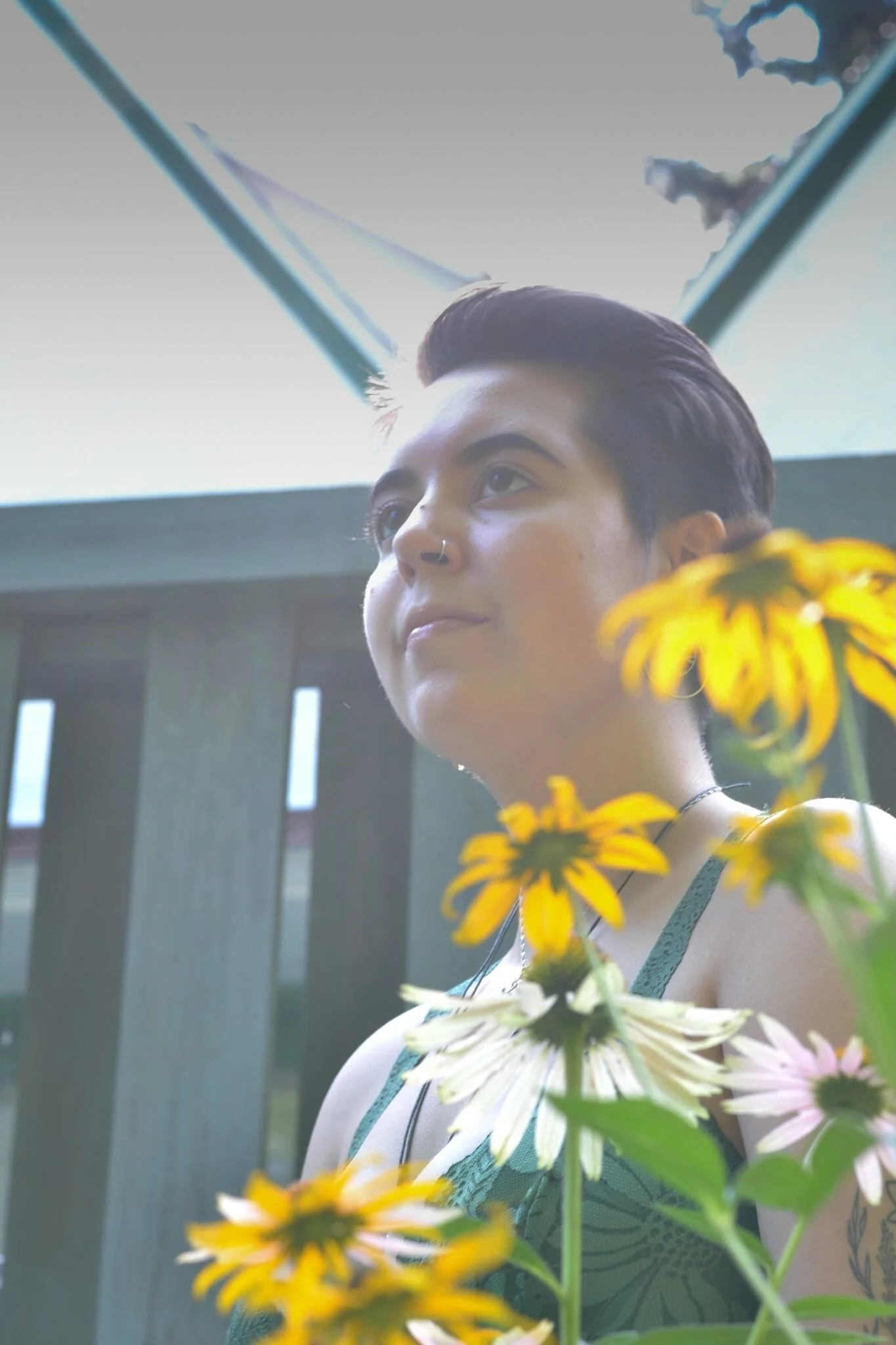 Jo, a white person with short brain hair, is sitting in front of a blue gate with yellow flowers in the foreground. They are staring serenely past the camera.