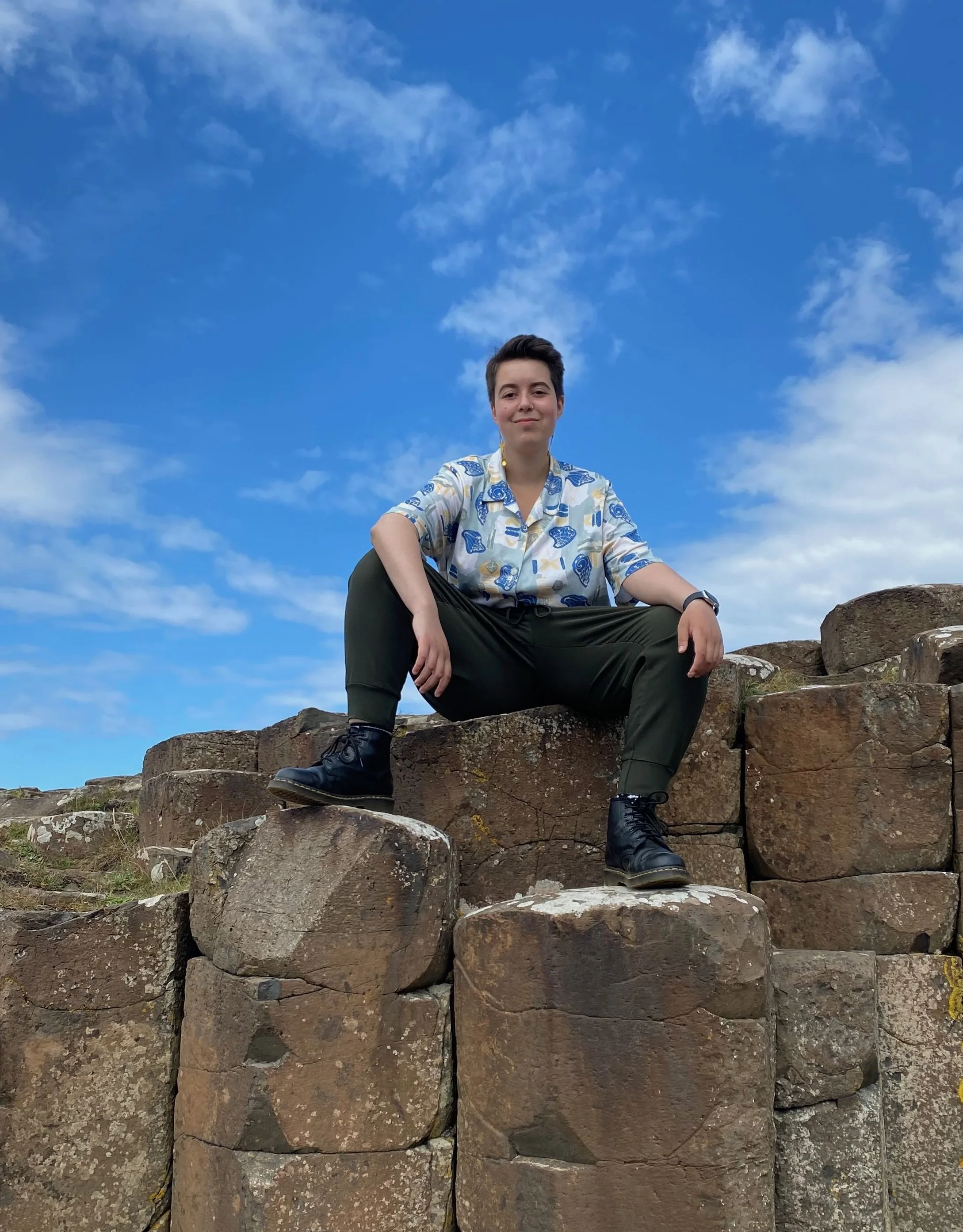 A photo of Jo, a white person with short brown hair, sitting on the hexagonal stones at the Giant's Causeway. Their legs are parallel with their shoulders and their arms are resting on their knees.