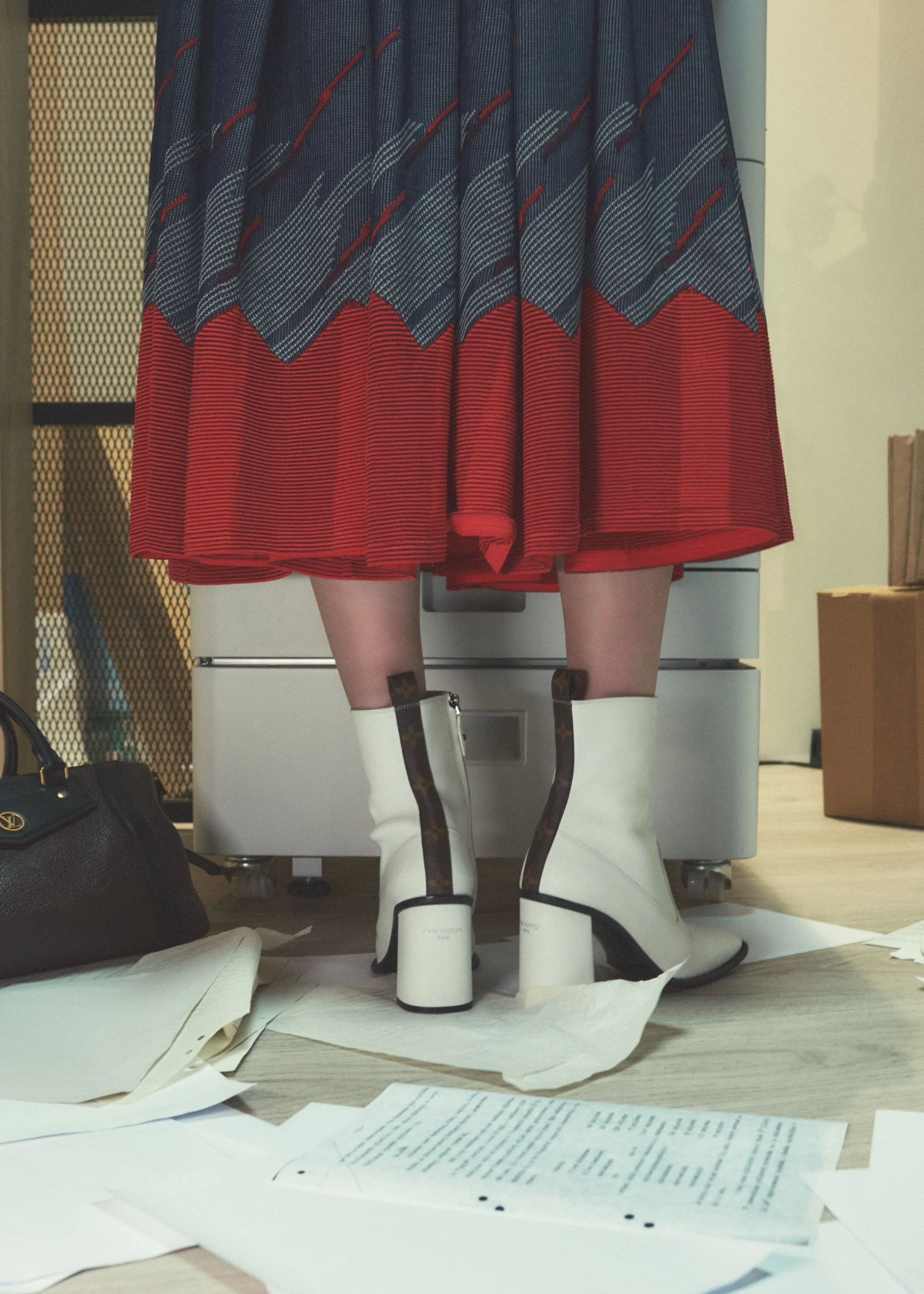Person wearing platform white boots with brown and gold details, red and plaid skirt, standing on scattered papers and near a brown handbag in a room with light-colored walls and flooring.