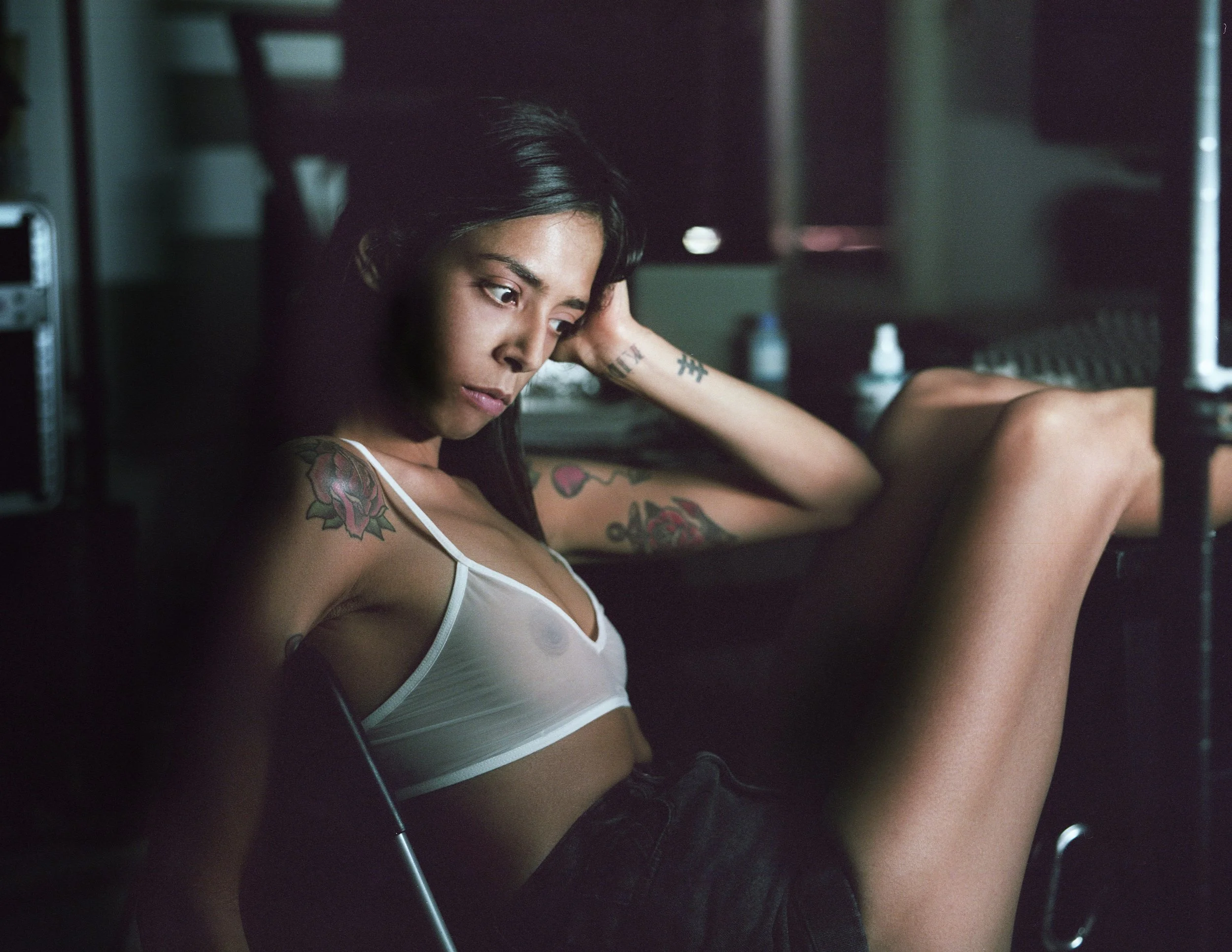 A woman with tattoos and wearing a white sheer top, sitting in a dark room looking contemplative.
