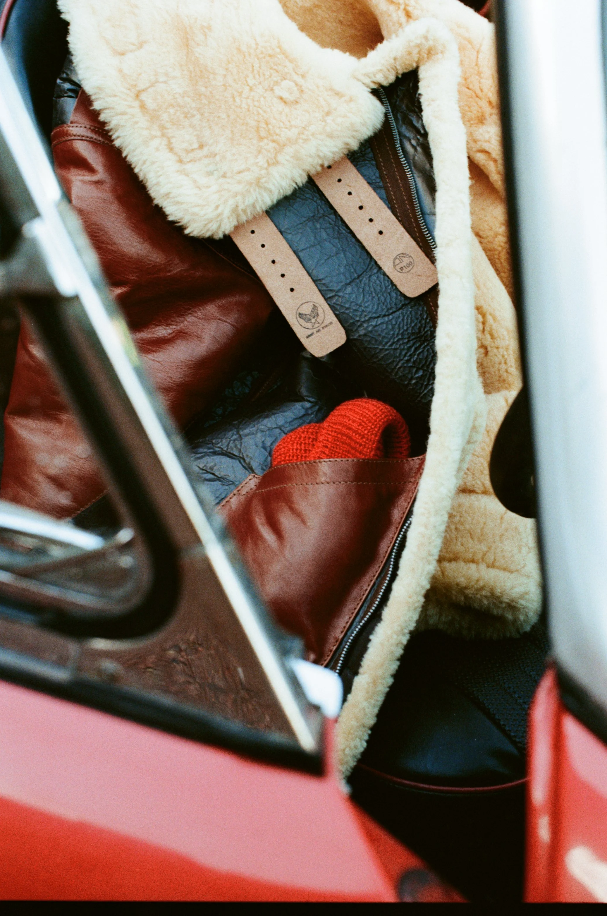 Open glove compartment in red vehicle containing a red knit hat, a belt, a black pouch, and a beige fleece jacket.