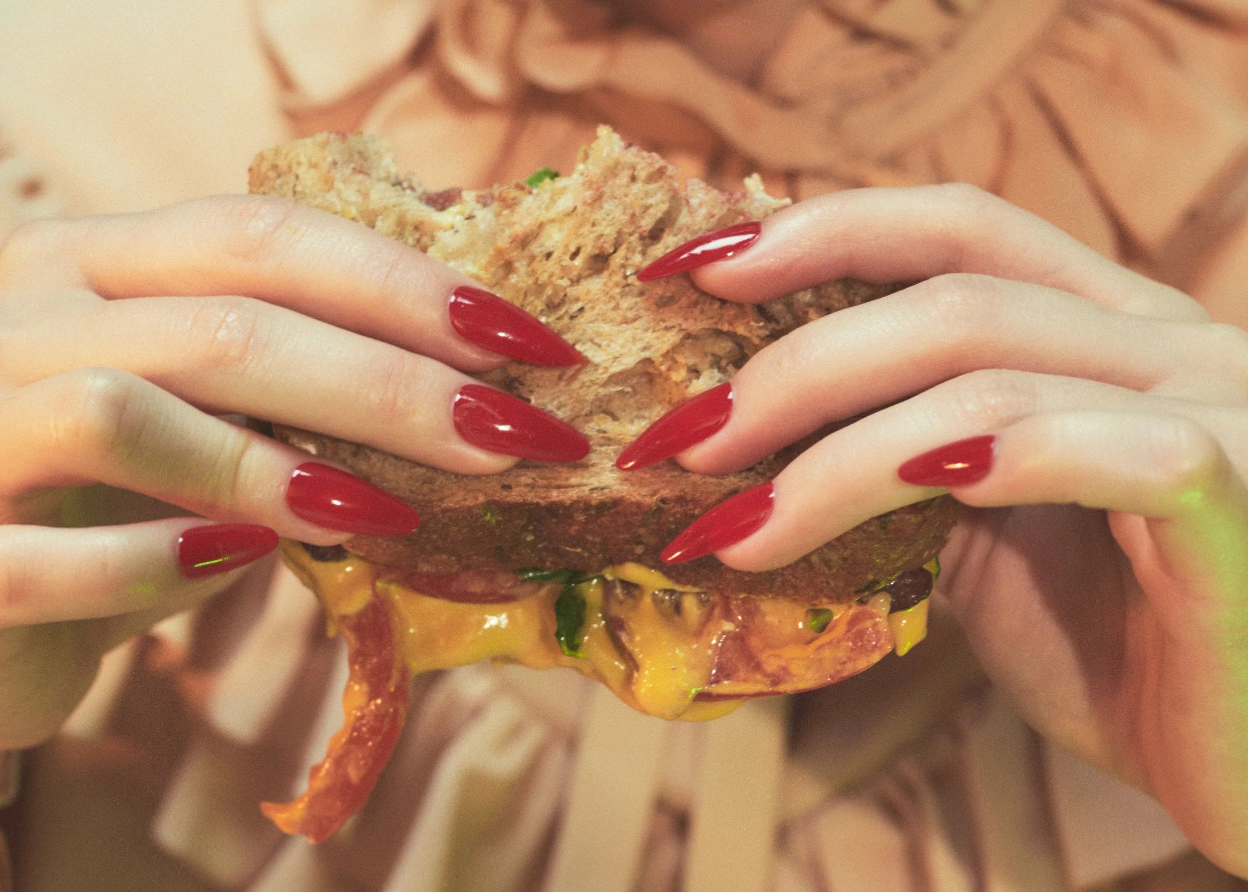 Person with long red painted nails holding a large, partially eaten sandwich with bread, meat, cheese, vegetables, and condiments.