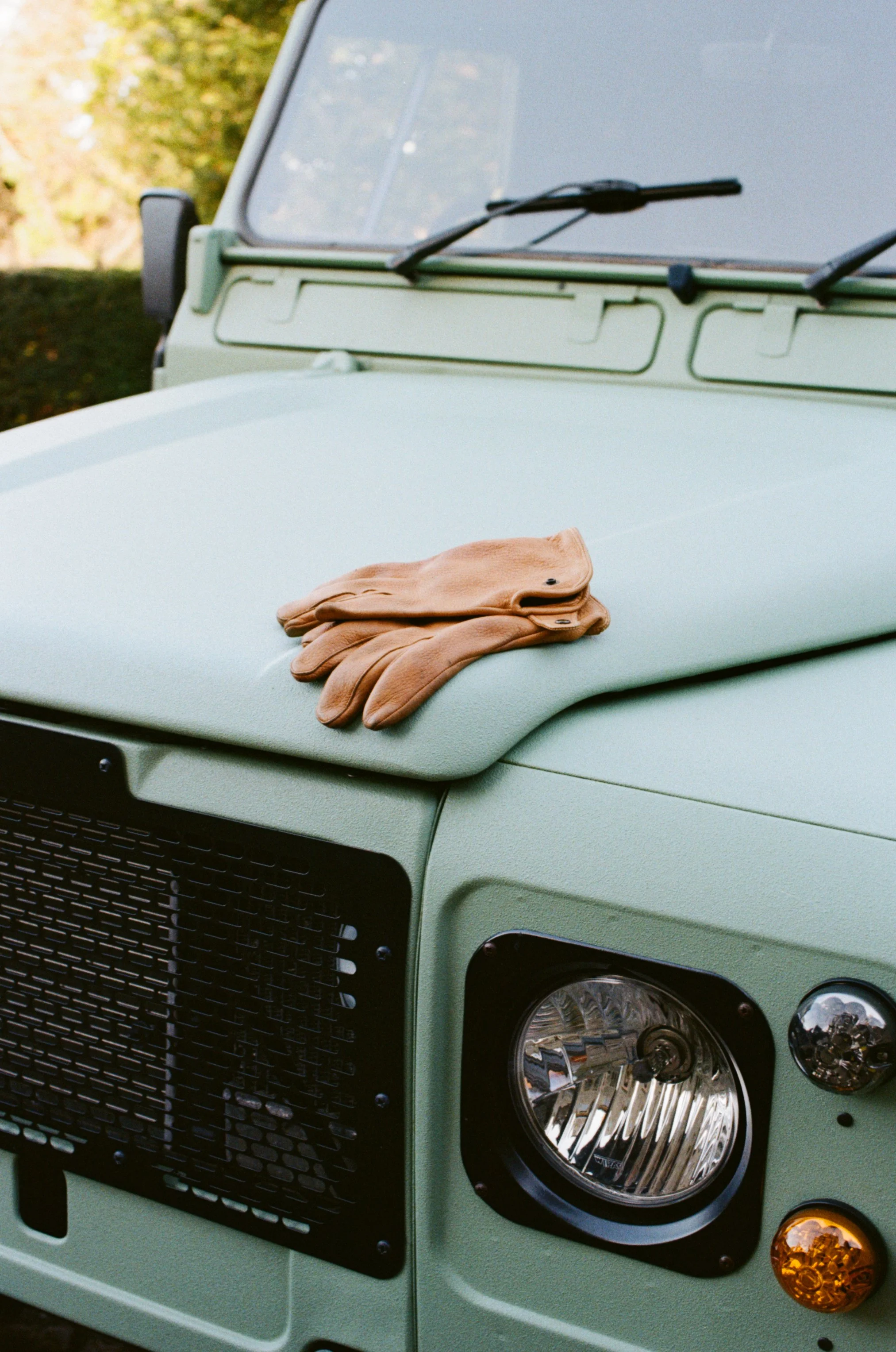 A pair of brown leather work gloves placed on the hood of a light green vehicle, near the headlight.