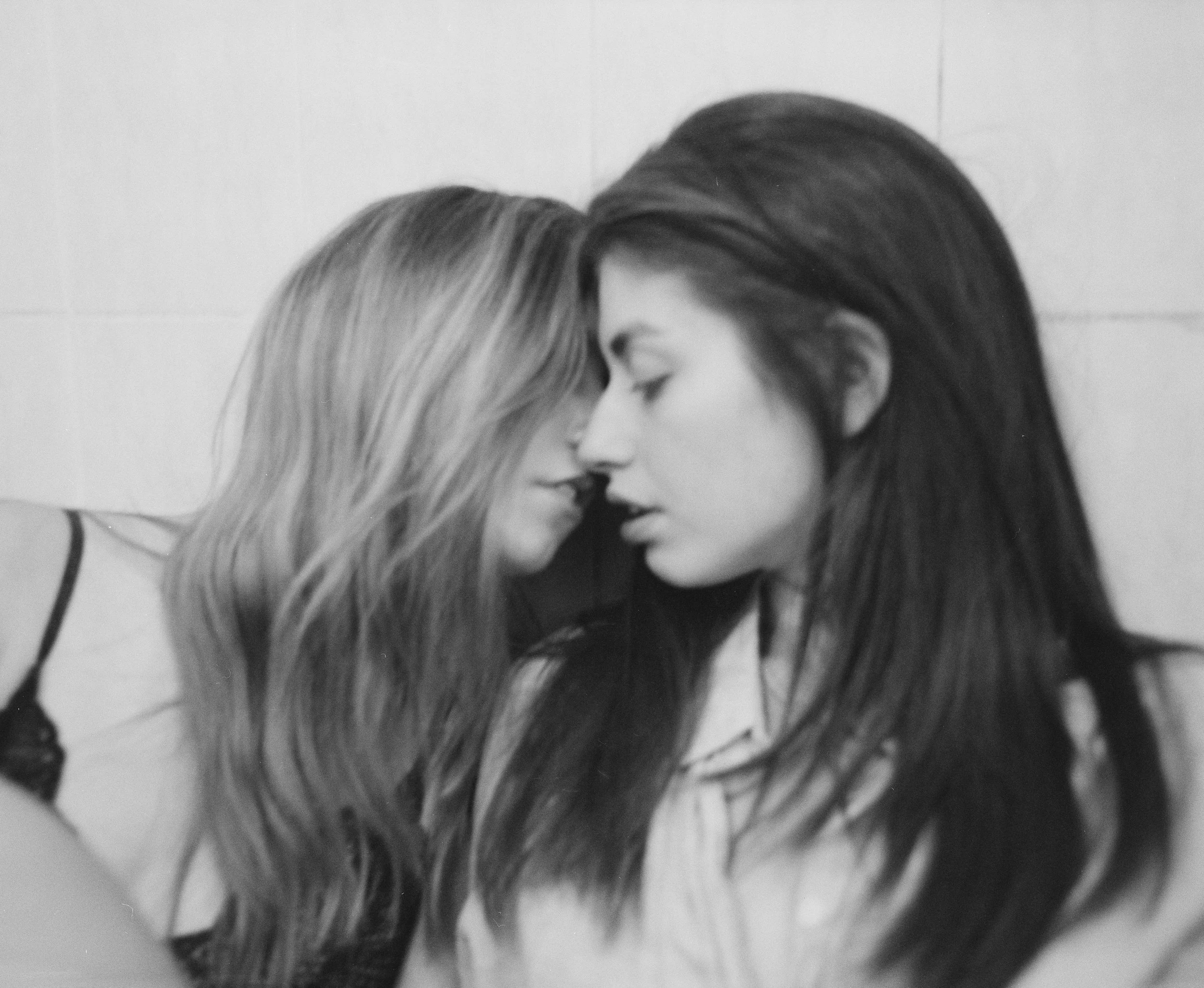 Two women with long hair gently touching foreheads, eyes closed, in an intimate pose against a plain background.