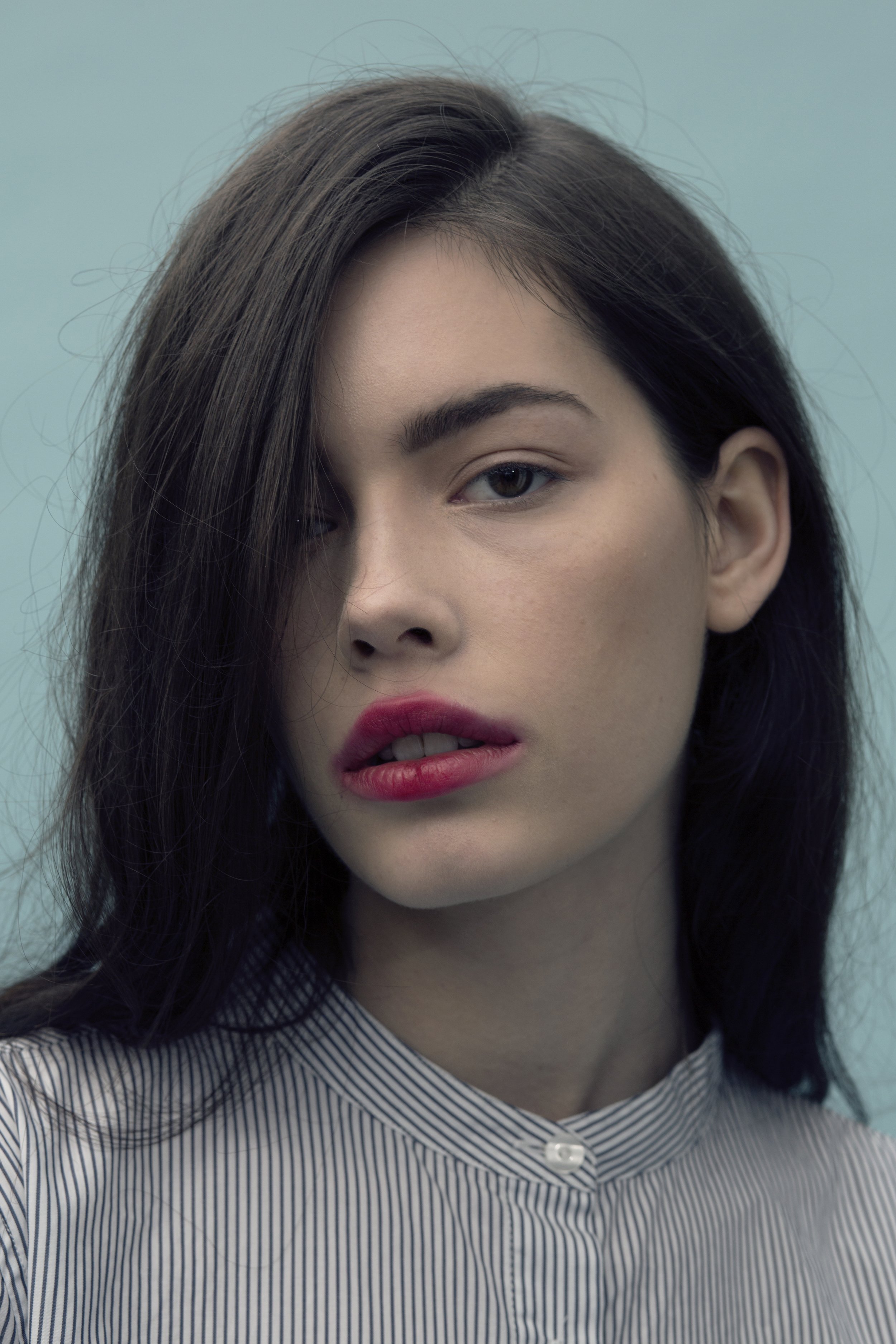 A close-up portrait of a young woman with dark hair, light skin, and makeup, wearing a striped shirt against a light blue background.