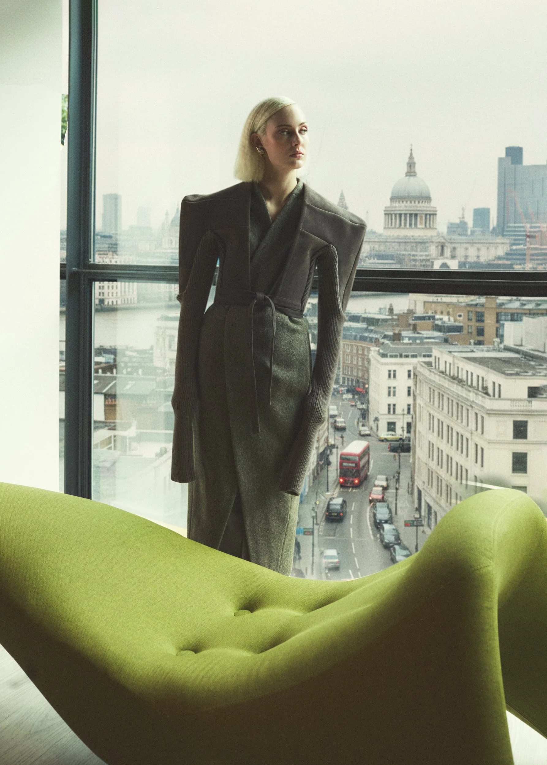 A woman with blonde hair standing inside an office, looking out the large window at a cityscape including the London St. Paul's Cathedral, with a green modern lounge chair in the foreground.