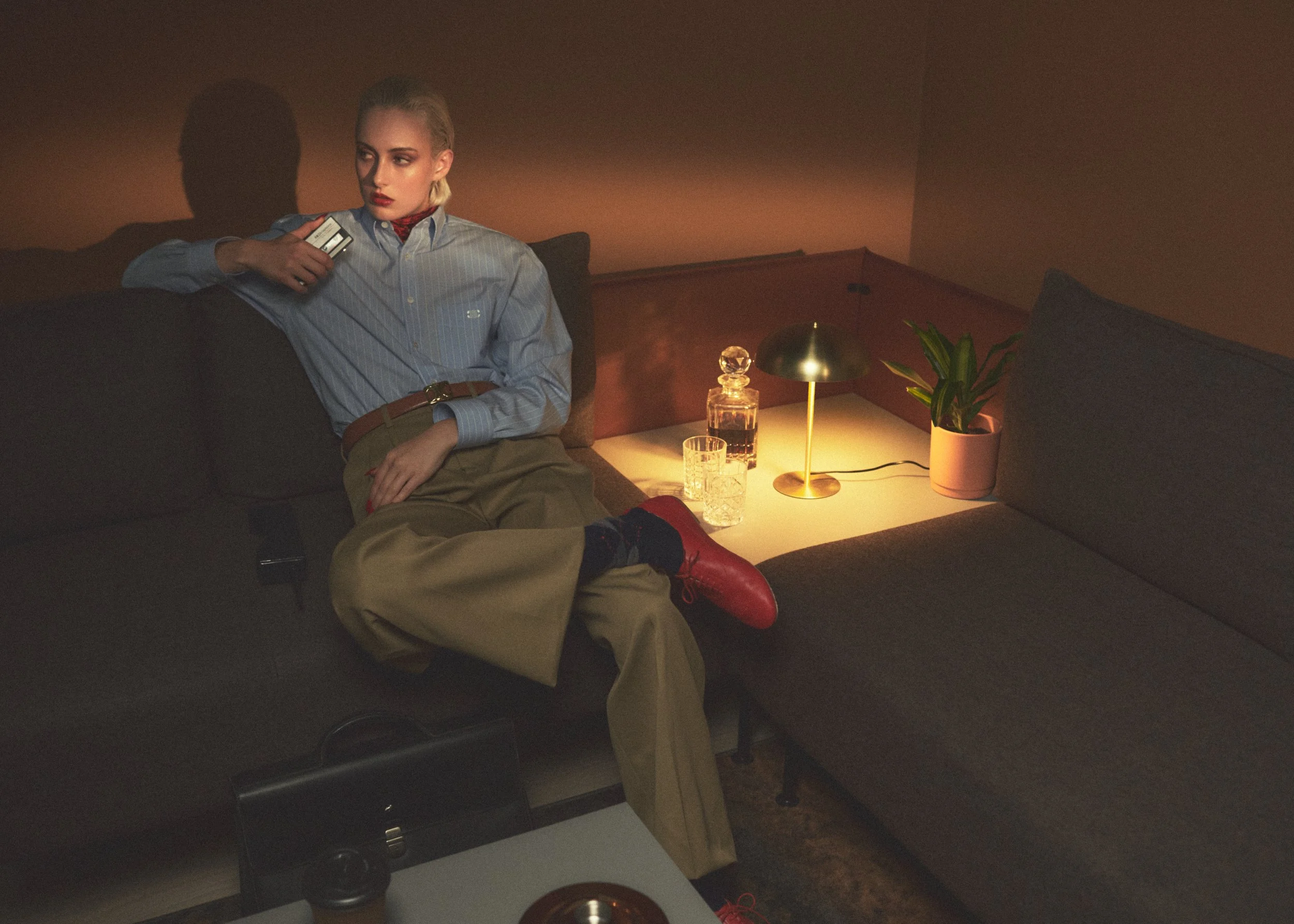 A woman with blonde hair sitting on a dark brown sofa in a dimly lit room. She is wearing a light blue striped shirt, khaki pants, and red shoes, with her legs crossed and holding a phone in her right hand. There is a side table with a gold lamp, a p