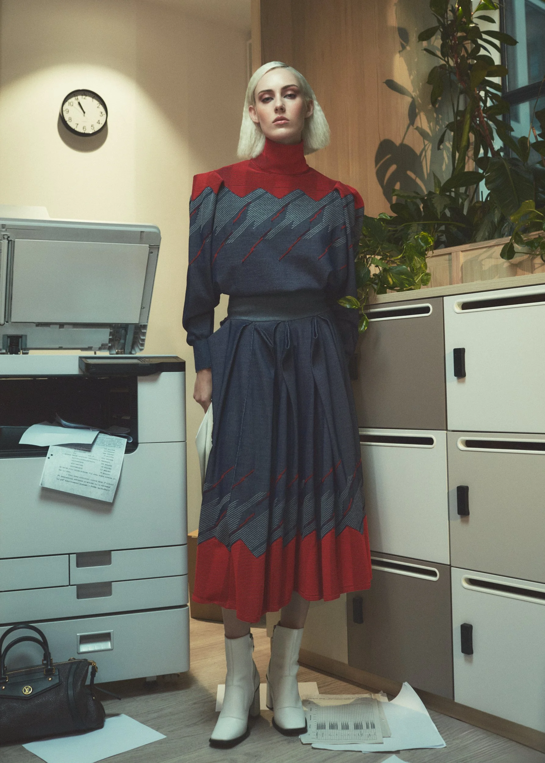 A woman with platinum blonde hair and fair skin stands in an office, wearing a long pleated skirt and a colorful sweater with a geometric pattern, white boots, and holding a white clutch bag. There is a potted plant and office furniture behind her, a
