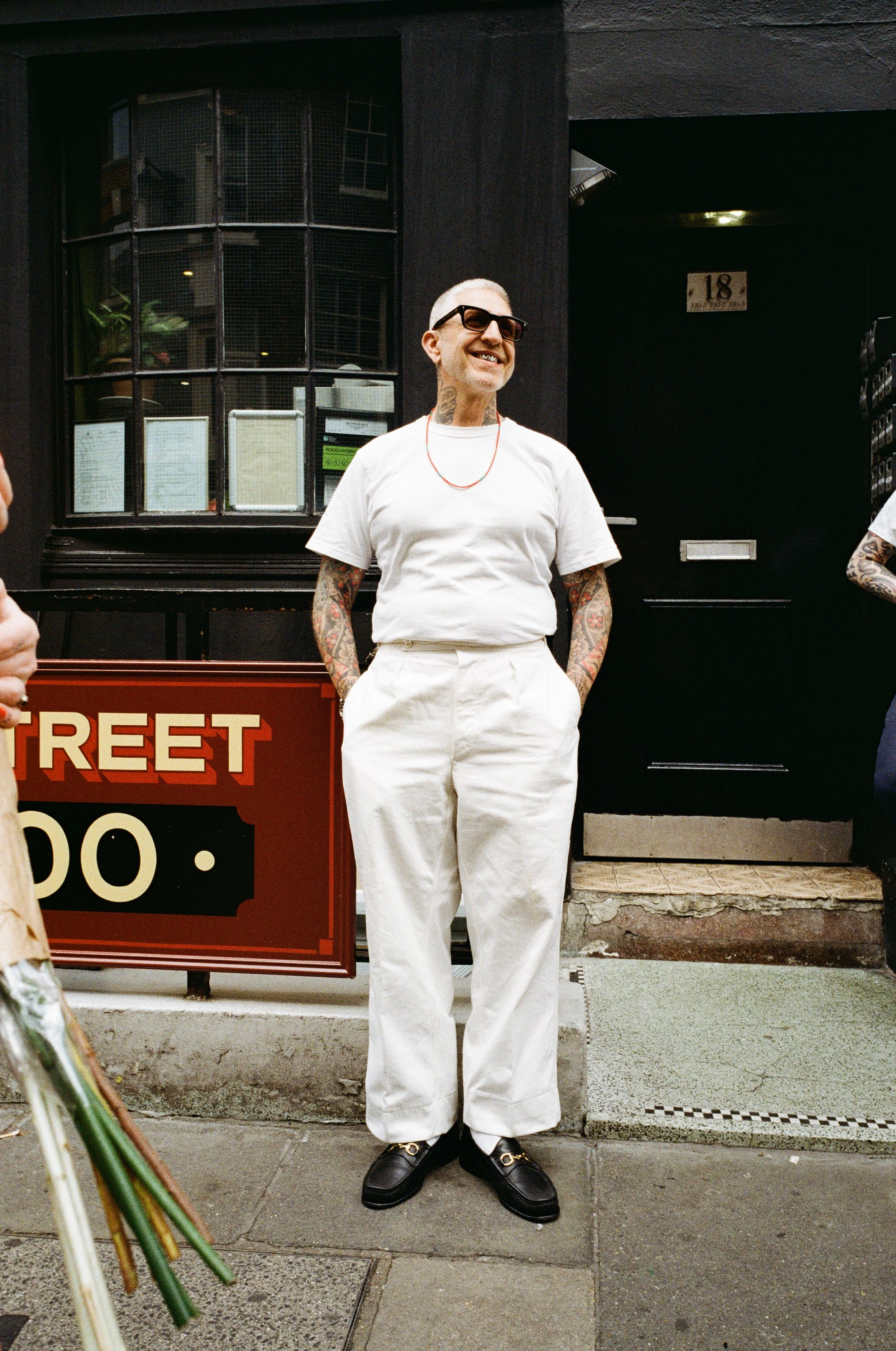A man with tattoos, wearing glasses, a white t-shirt, and white pants, smiling and standing on a city sidewalk outside a building with a sign that reads 'STREET' in red letters.