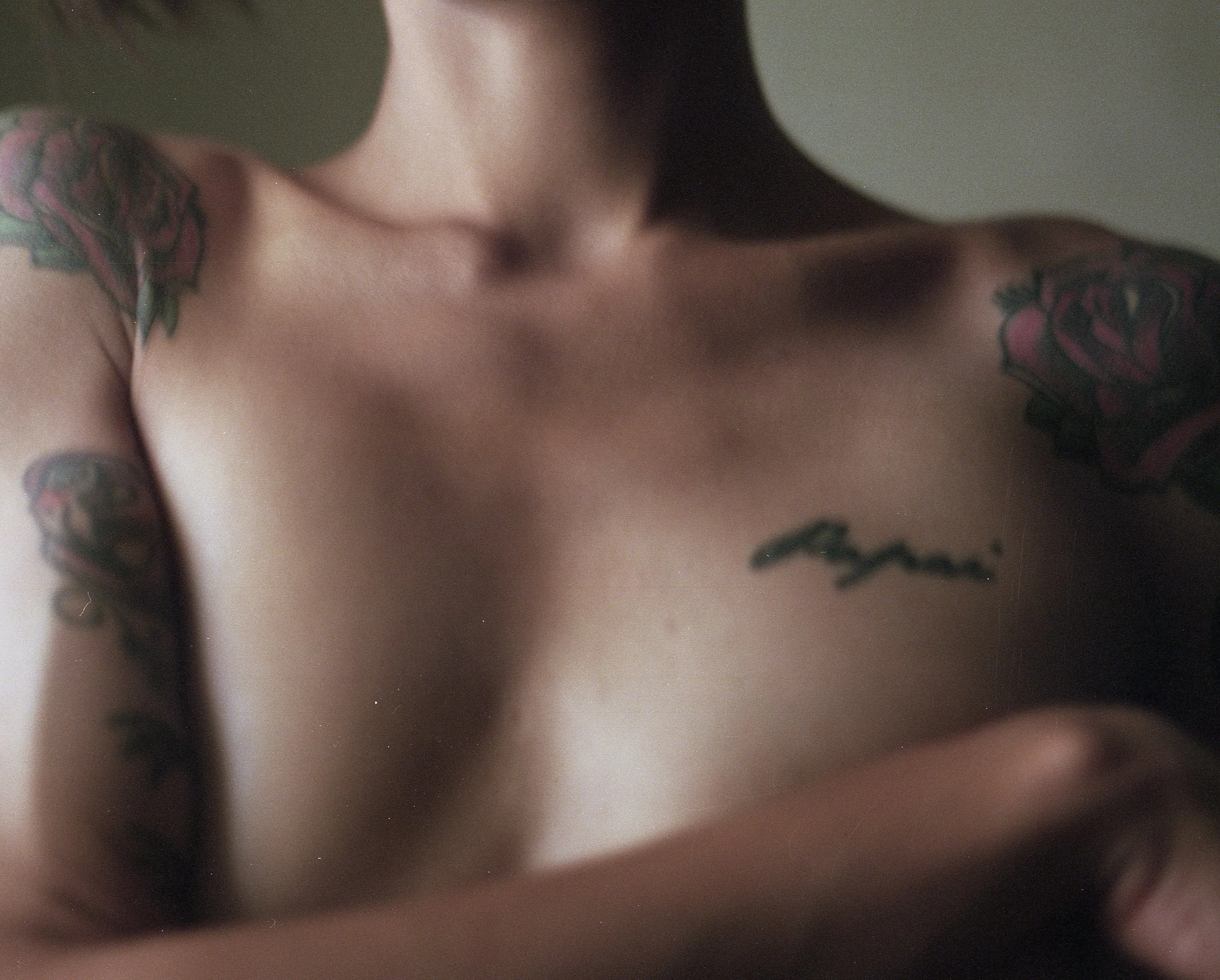 Partial view of a person's upper body showing tattoos on shoulders and chest, with the person's right arm crossed over their chest.