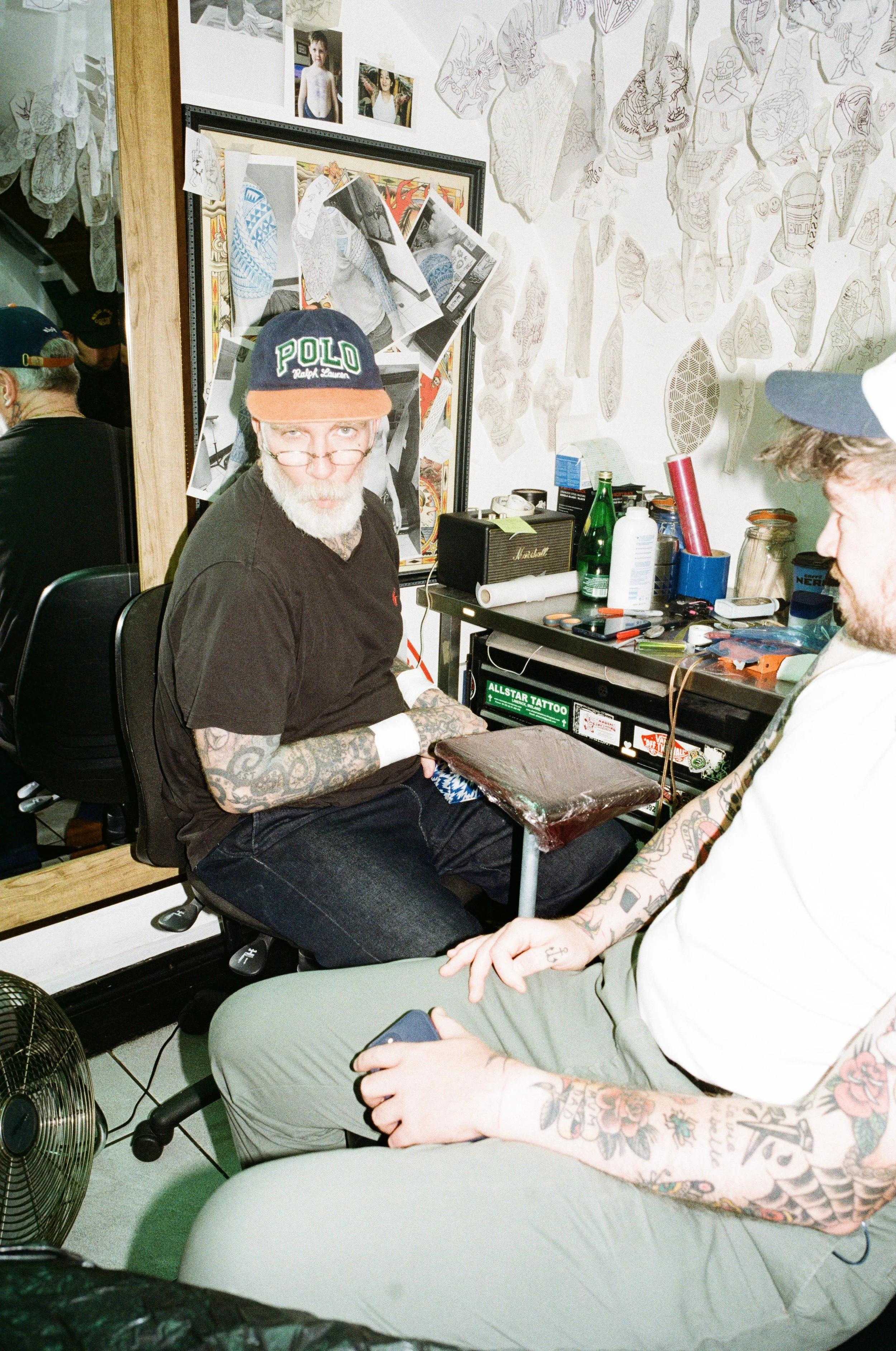An older man with tattoos on his arms and wearing a black Polo cap is sitting in a tattoo parlor. He is looking at the camera, with a partly visible younger person with tattoos on their arms sitting nearby, holding a phone. The room has a workbench w