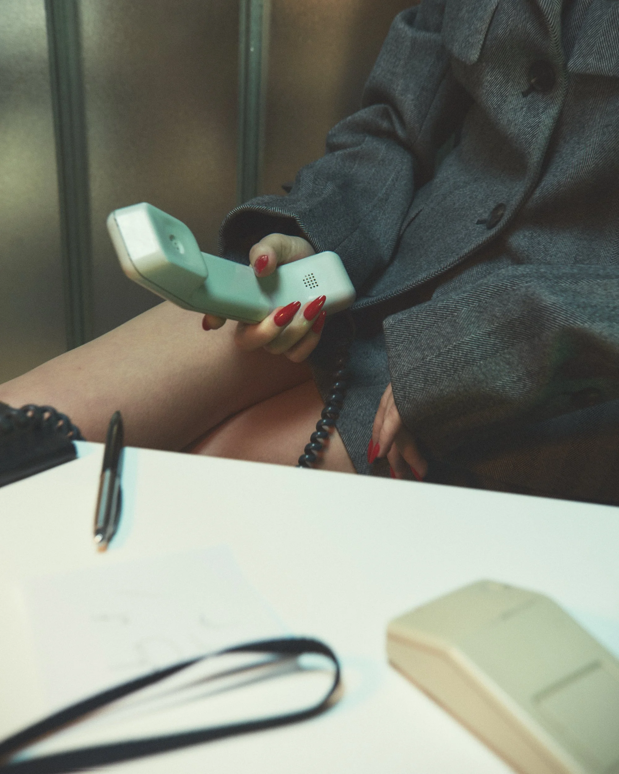 A person with red-painted nails holding a vintage landline phone, seated at a desk with a pen, paper, and calculator visible.