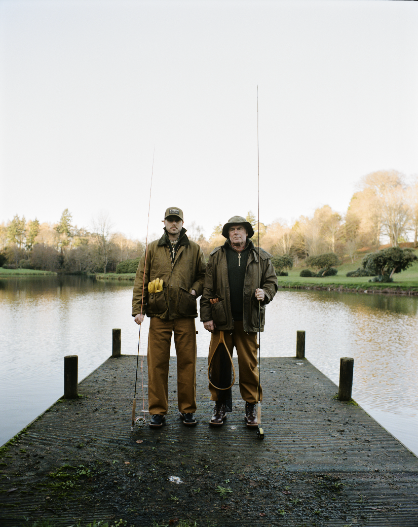 Two men standing on a wooden dock by a lake, holding fishing rods, wearing outdoor fishing gear, with trees in the background.