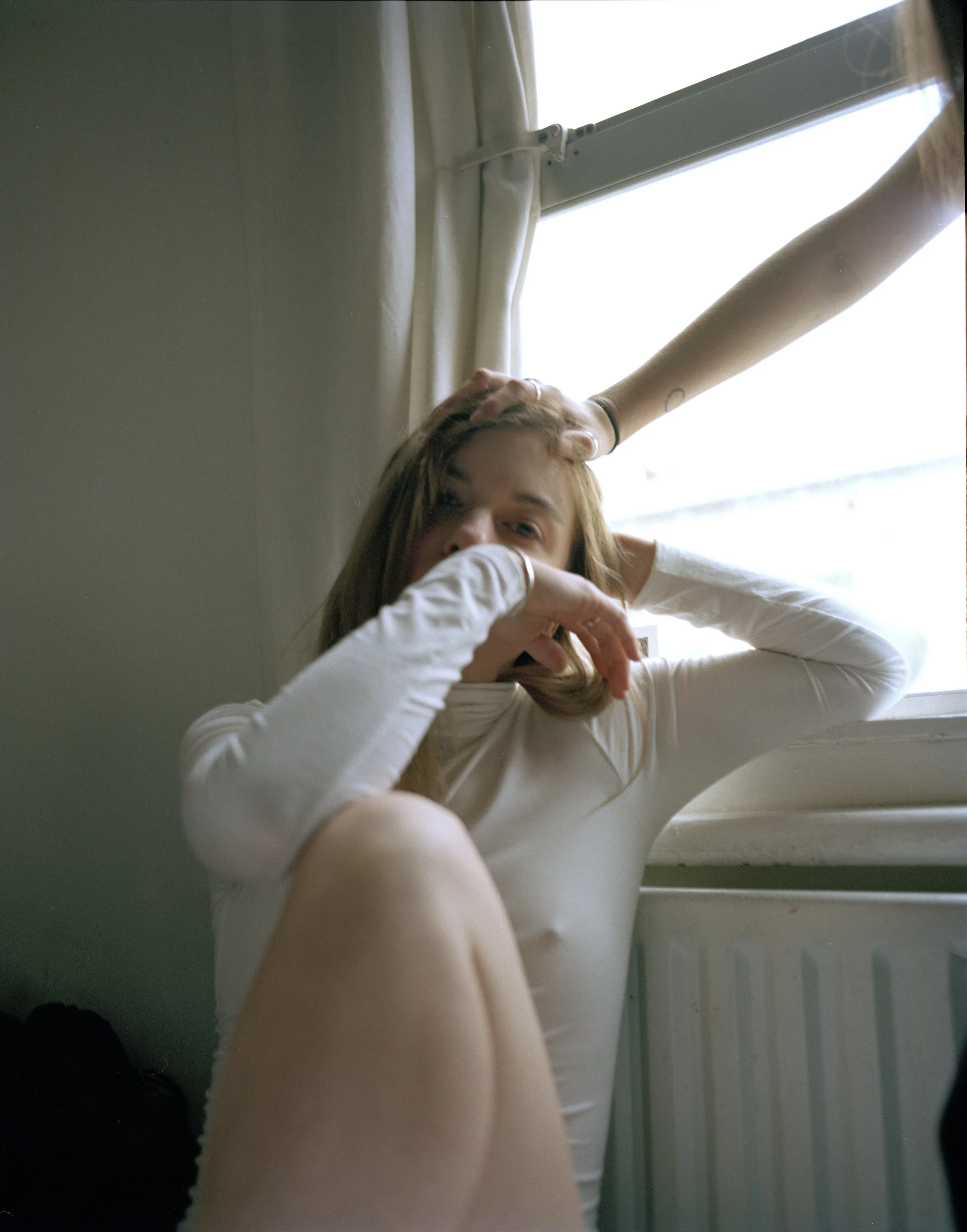 A young woman sitting on the floor near a window, with one hand on her head and the other covering her mouth, looking at the camera. A person's hand is touching her head. There's a radiator under the window and curtains tied to the side.