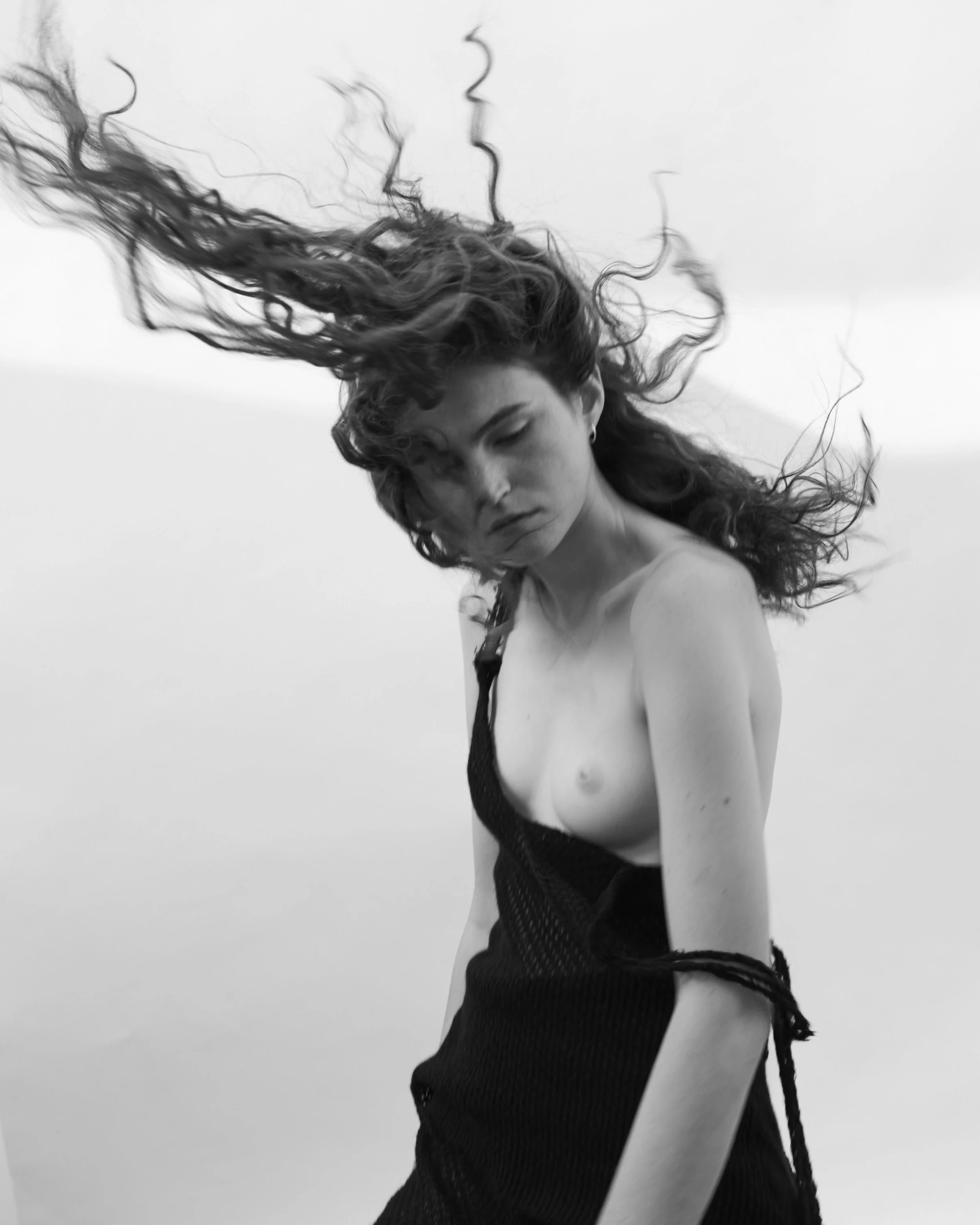 A black and white photo of a woman with long, curly hair blowing in the wind, wearing a loose black top off her shoulder, with a neutral expression.