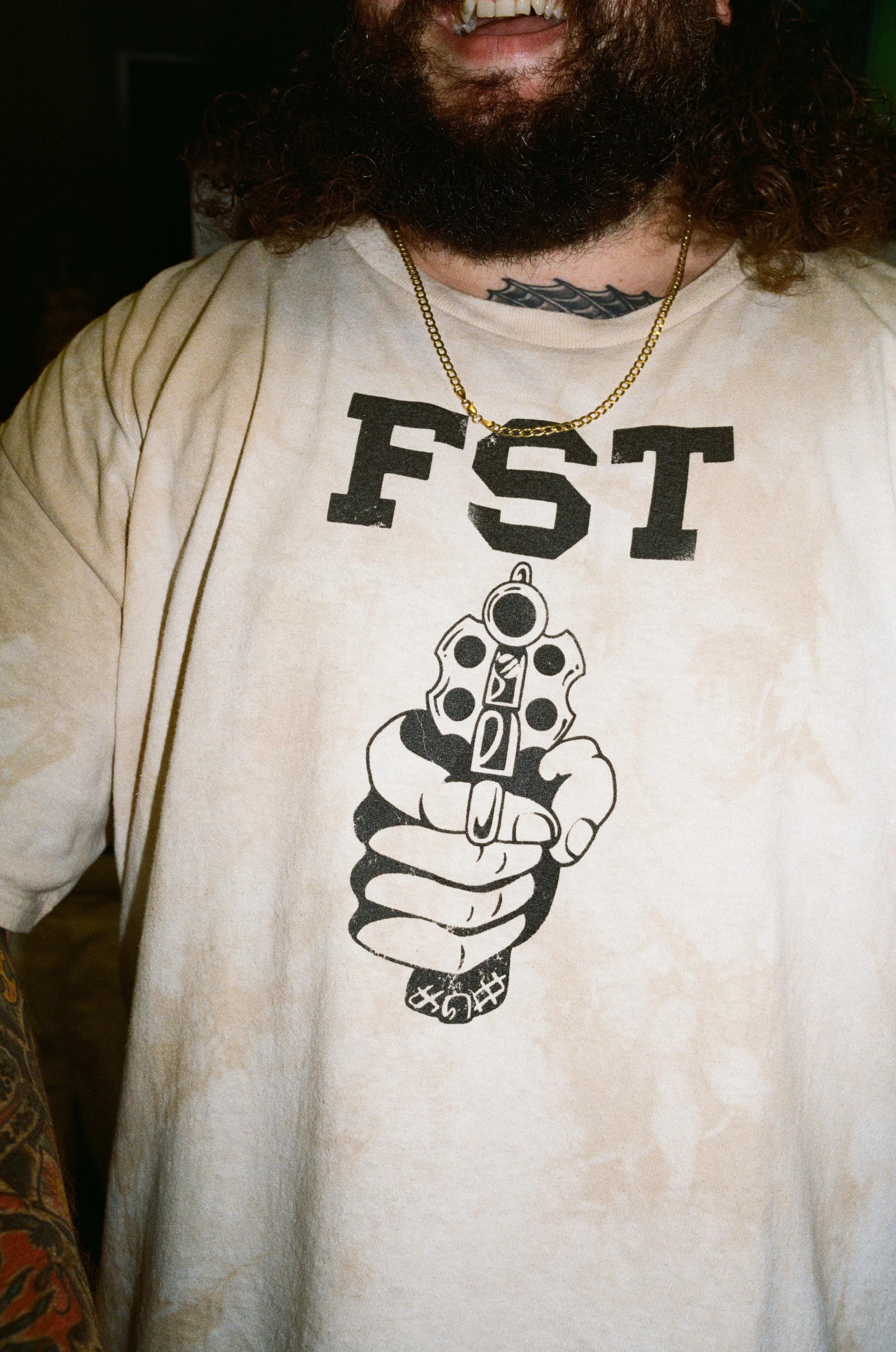 A person wearing a beige t-shirt with a graphic of a hand holding a gun and the letters 'FST' above it. The person is also wearing a gold chain necklace.