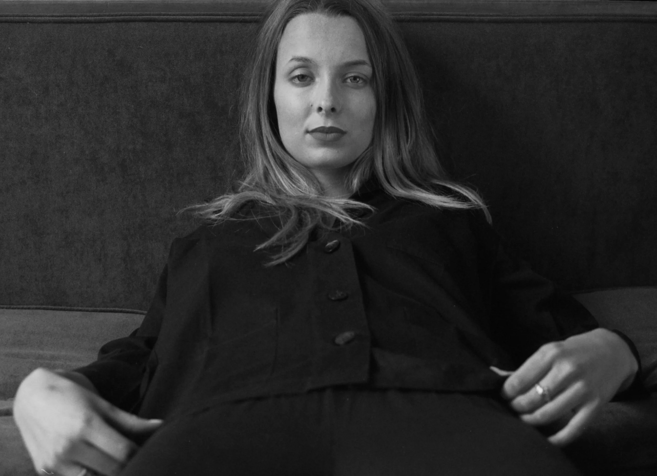 A black and white photo of a woman with long hair, sitting on a couch with a dark, textured wall behind her. She is wearing a dark jacket with buttons and looking directly at the camera.